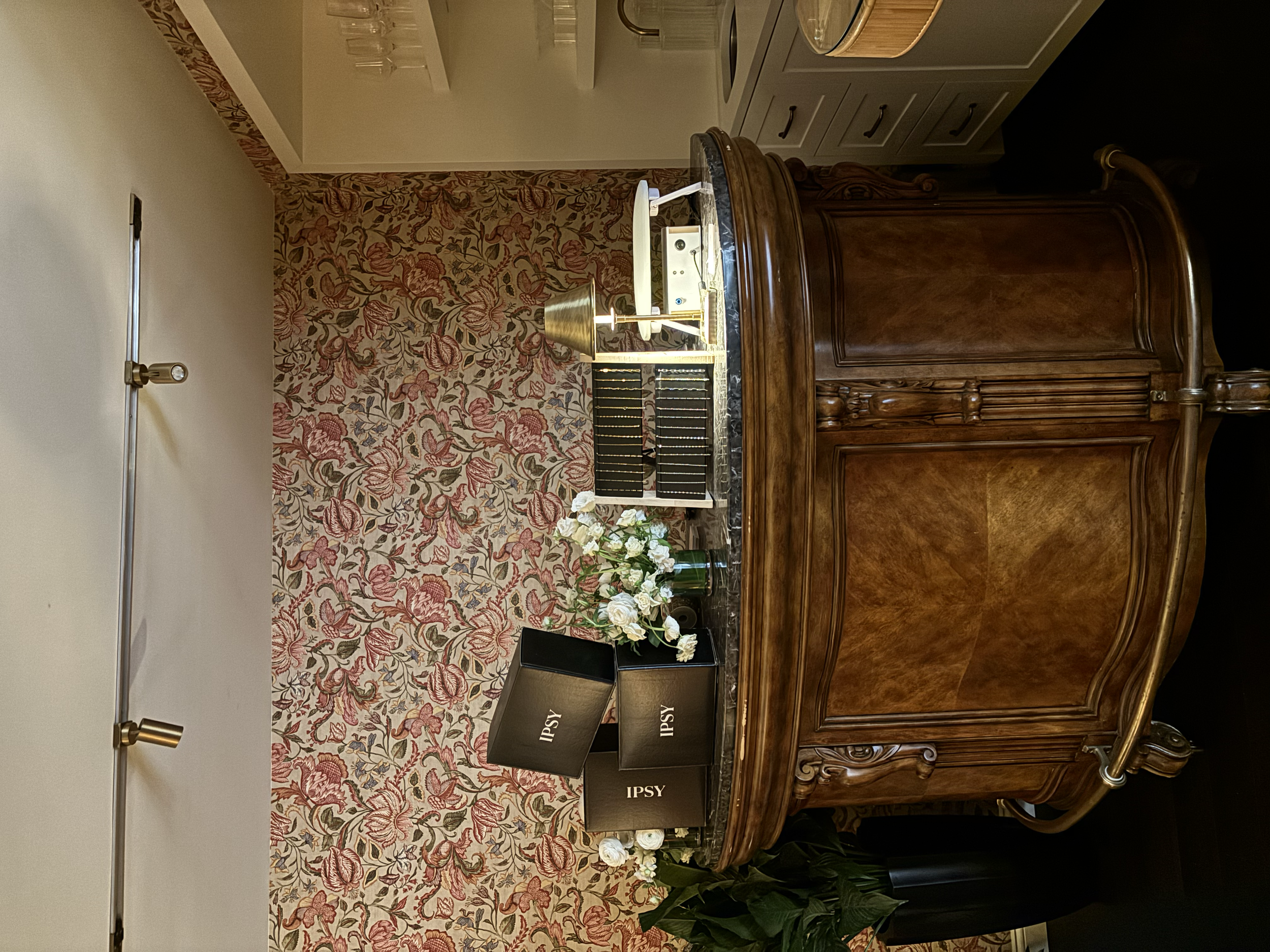A wooden bar with a marble top in a room with floral wallpaper, books, a flower arrangement, a table lamp, and glasses on a shelf.