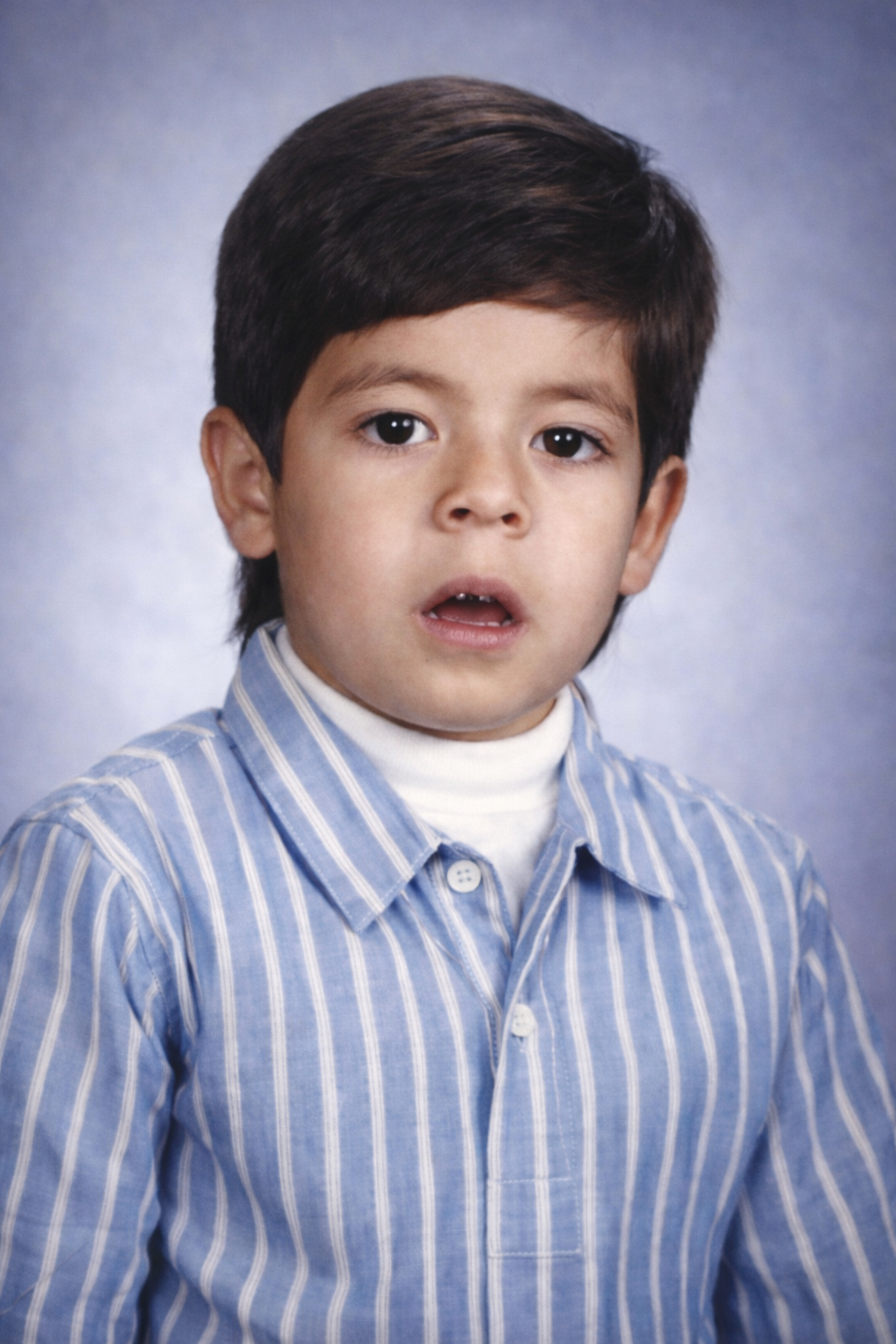 A young boy with dark hair, wearing a blue and white striped shirt and a white turtleneck, has a surprised or confused expression on his face against a blue gradient background.