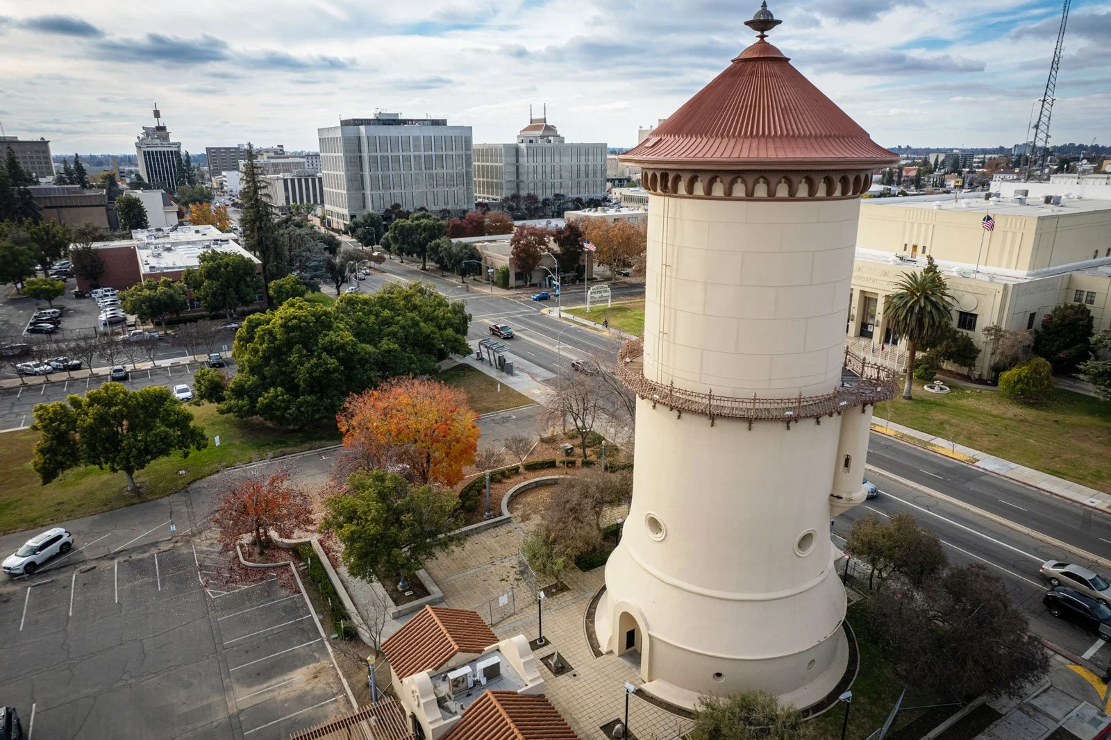 An aerial view of a cityscape featuring a tall, white cylindrical tower with a reddish-brown conical roof. Surrounding the tower are green trees, some with autumn colors, and paved walkways. In the background, there are various modern office buildings and a street with cars and traffic signals.