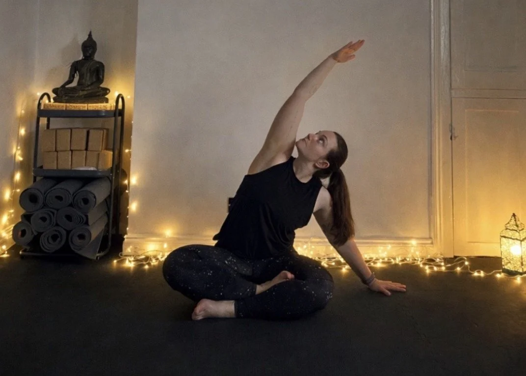 A woman practicing yoga indoors, sitting cross-legged on the floor, reaching upward with her right arm, surrounded by warm string lights and yoga mats.