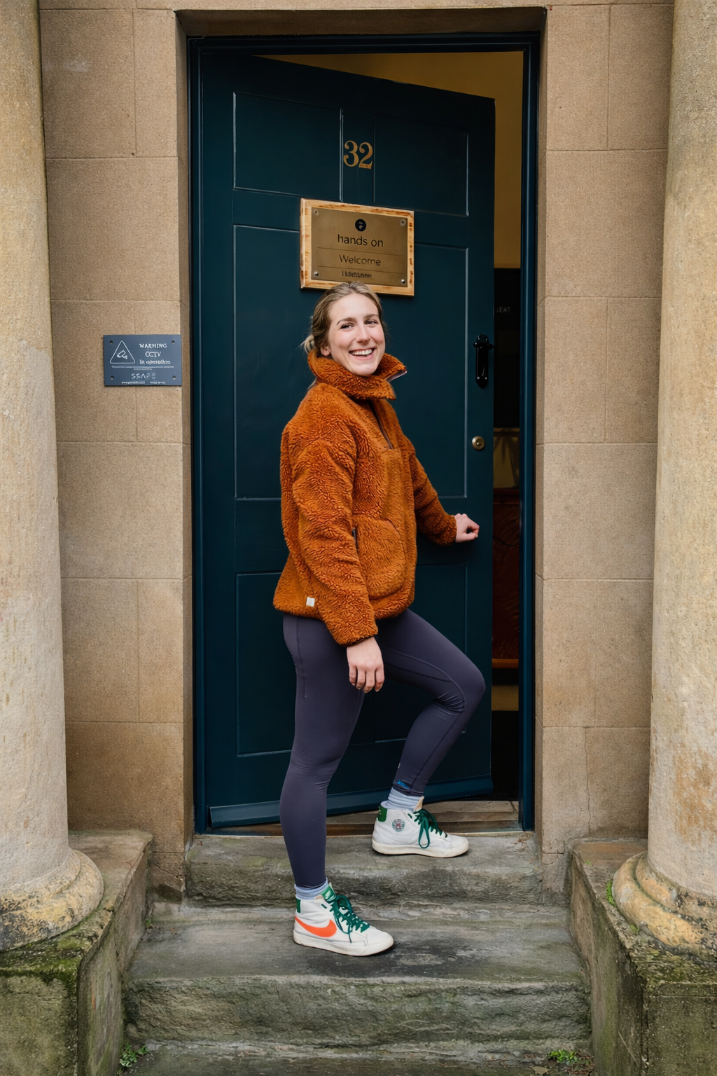 A woman standing at an open door, smiling, wearing a brown fleece jacket, gray leggings, and white sneakers with green and orange accents.