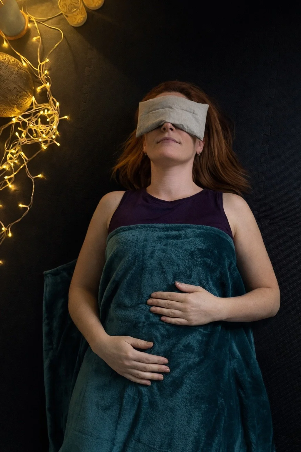 Woman lying down with an eye mask over her eyes, covered with a blanket, on a black surface, with a string of fairy lights and candles nearby.