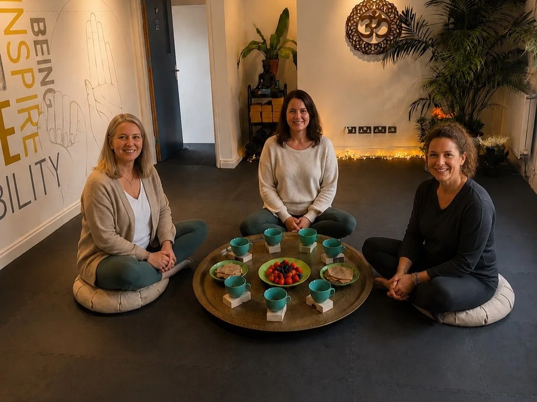 Three women sitting cross-legged on cushions around a tray with green cups and a bowl of strawberries, inside a room with plants and a wall with a large OM symbol.