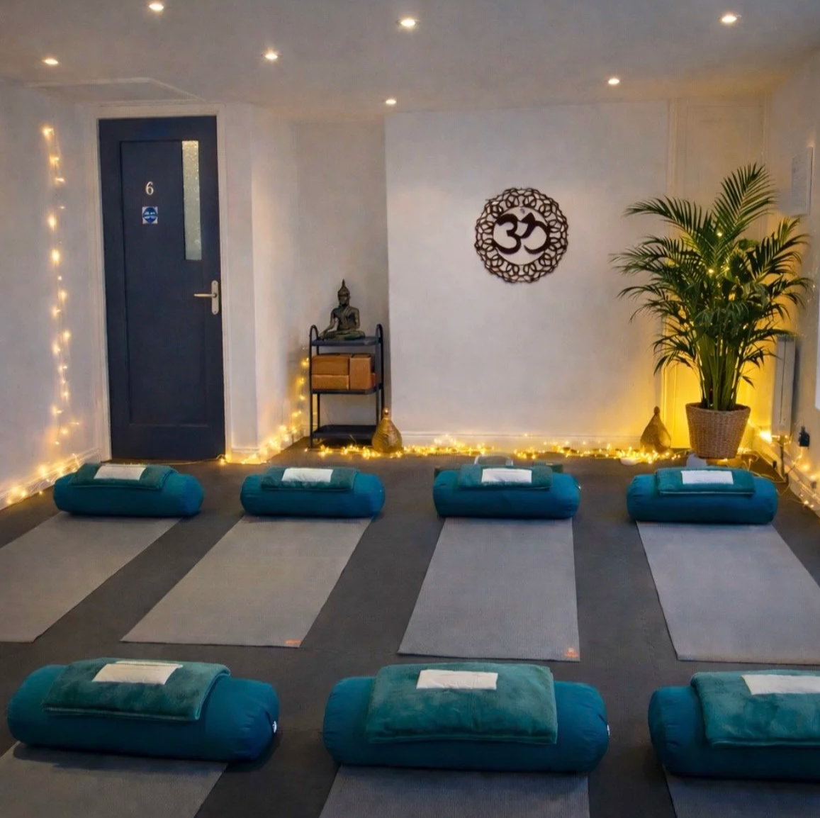 A meditation room with eight teal bolster cushions and mats on a gray carpet, a white wall with a Buddha and Om symbol, string lights, a large potted plant, and a small shelf with a Buddha statue.
