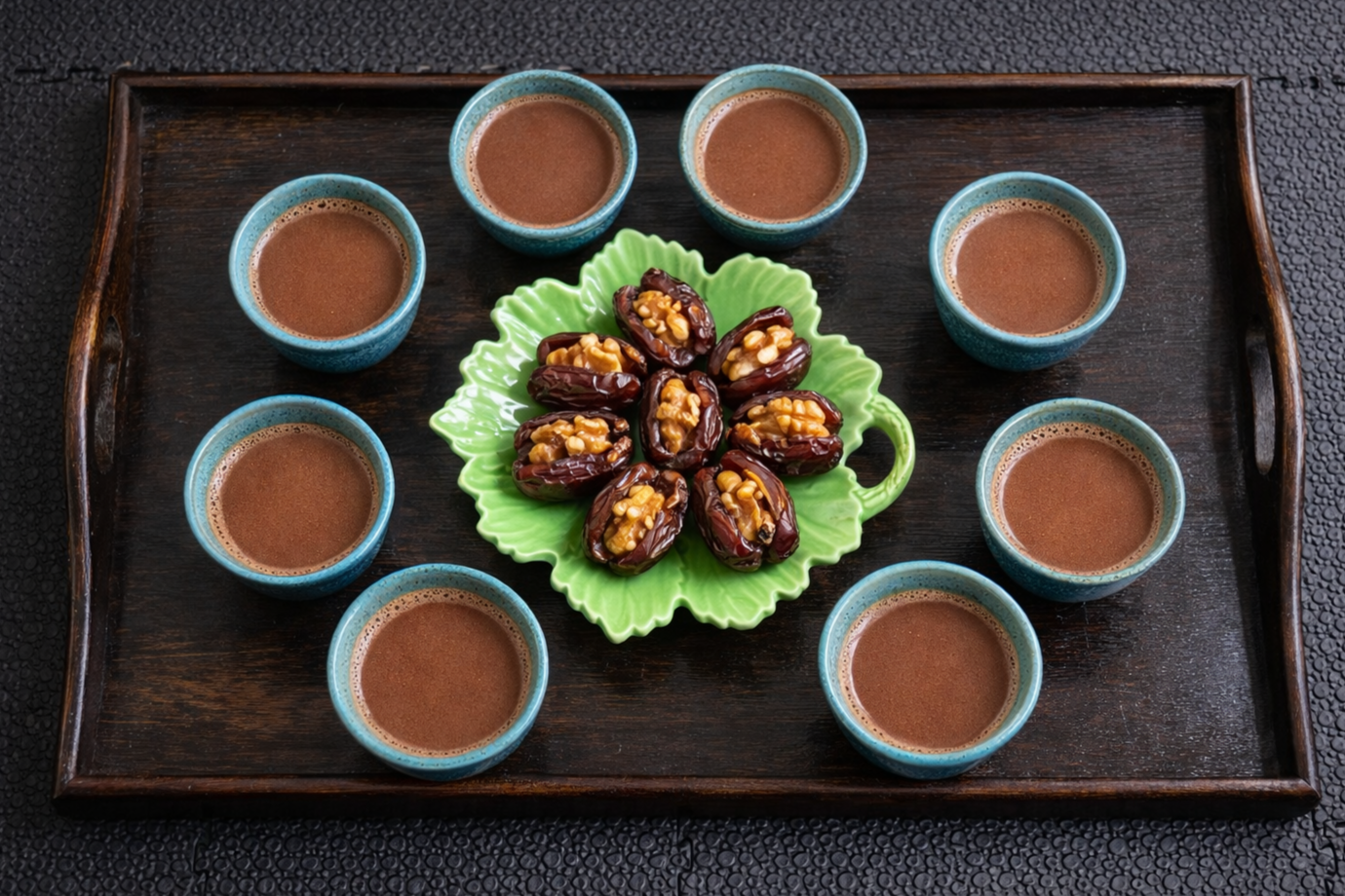 A wooden tray with eight small cups of cacao surrounding a green decorative plate filled with date and nut-filled desserts.
