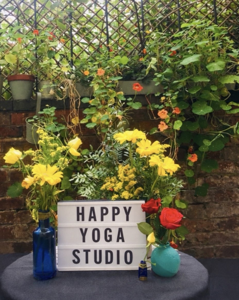 A decorative setup featuring a lightbox sign that reads 'HAPPY YOGA STUDIO', surrounded by vibrant yellow and red flowers in vases, with green foliage and garden plants in the background.