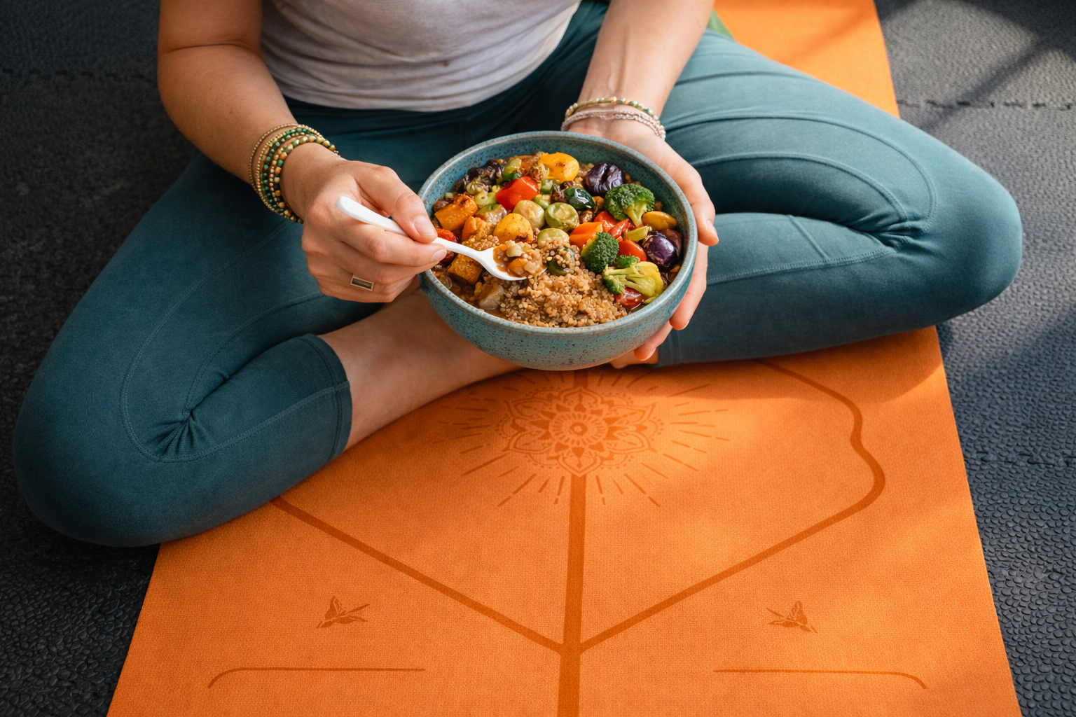 Person sitting cross-legged on an orange yoga mat, holding a bowl of mixed vegetables and grains, and eating with a white spoon.
