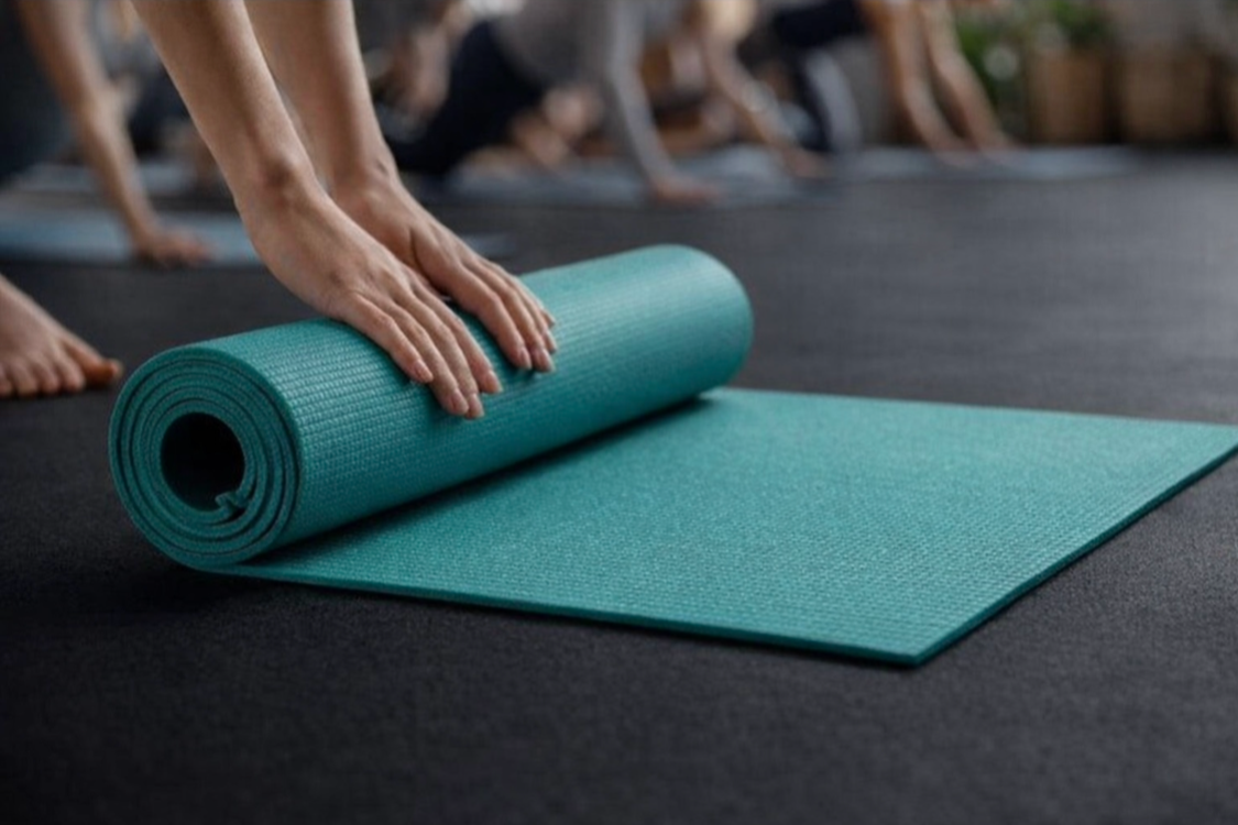 Person unrolling a turquoise yoga mat on a black floor during a yoga class.
