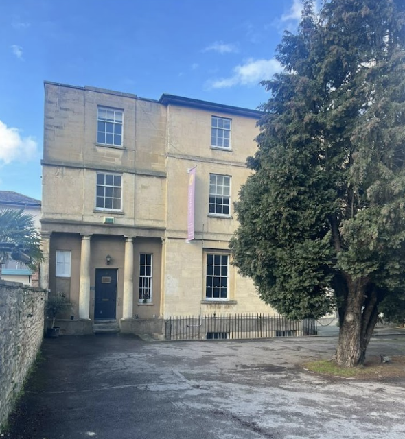 A three-story historic stone building with evenly spaced windows, a doorway with columns, a large tree to the right, and a paved driveway or parking area in front, under a blue sky with some clouds.