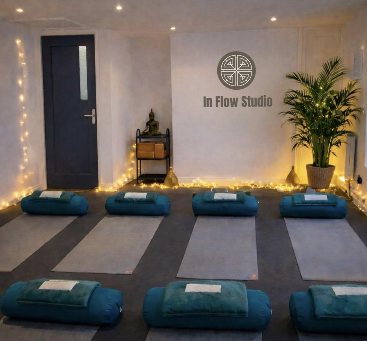 Yoga mats with bolsters and towels arranged for meditation in a softly lit studio with a Buddha statue, a potted plant, and a sign that reads 'In Flow Studio'.