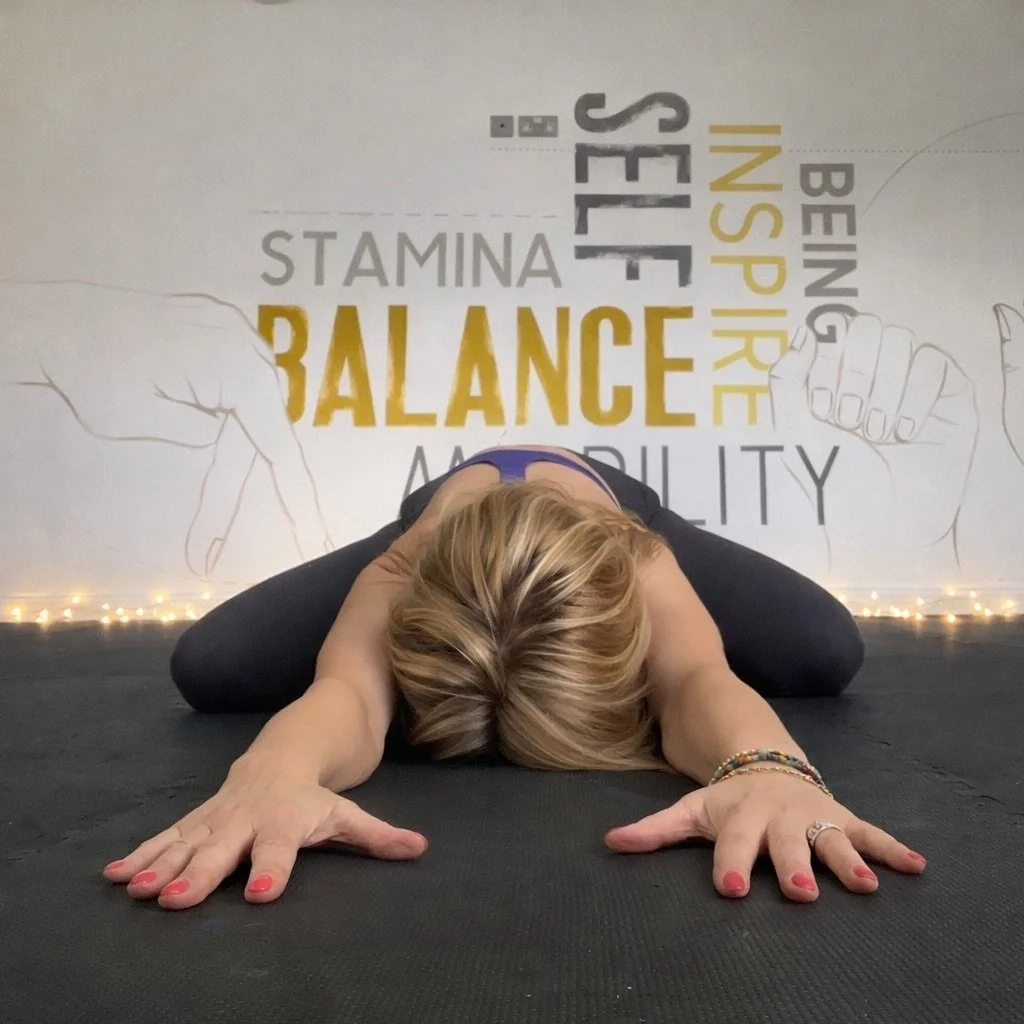 A woman in a yoga pose with her head down and arms extended forward on a black mat in a fitness studio with wall art featuring words like balance, self, independence, stamina, and flexibility.