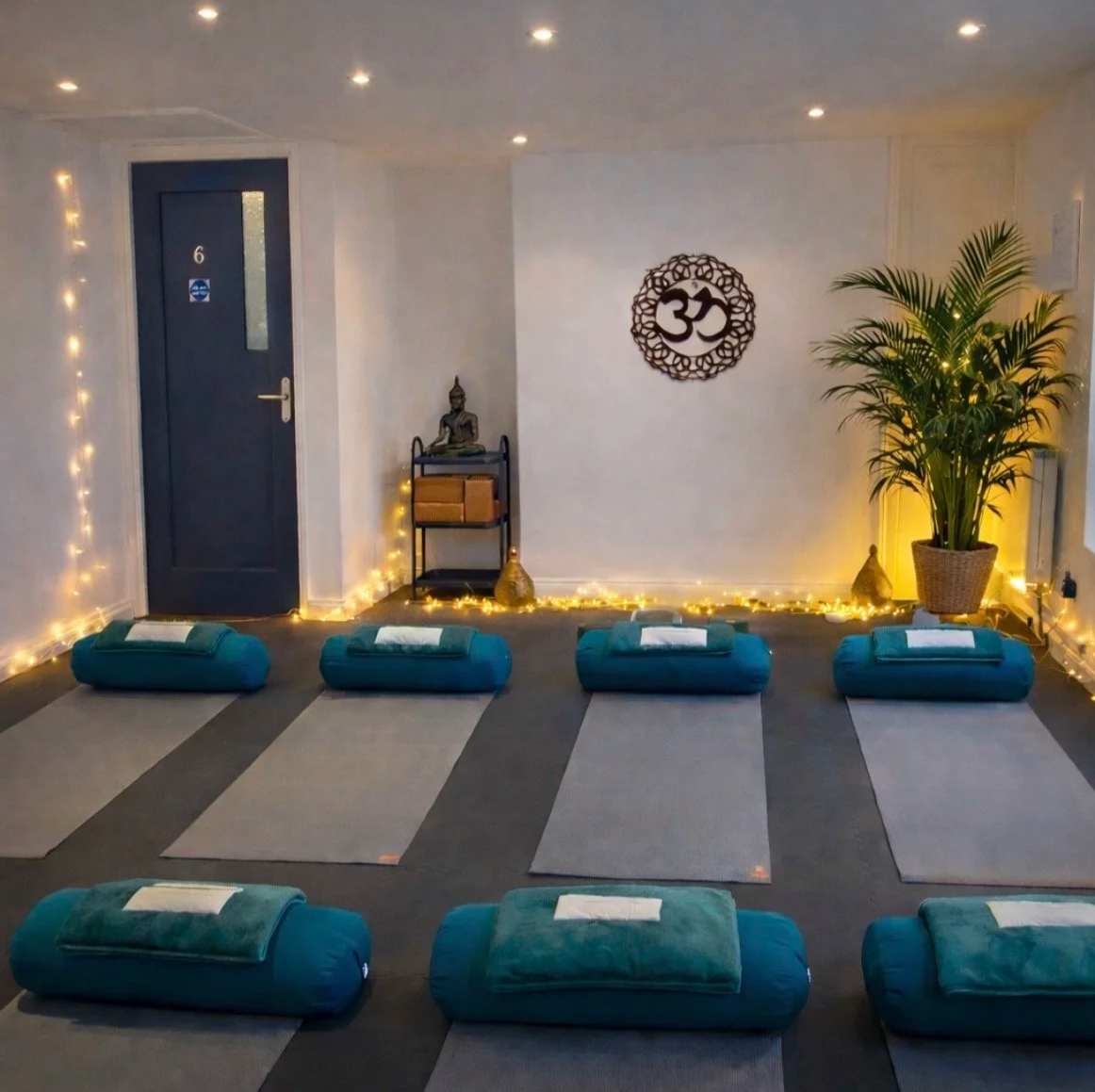 A serene yoga or meditation room with eight teal bolsters on gray mats, a Buddha statue, a leafy potted plant, and a large Hindu Om symbol on the wall. The room is decorated with warm fairy lights.