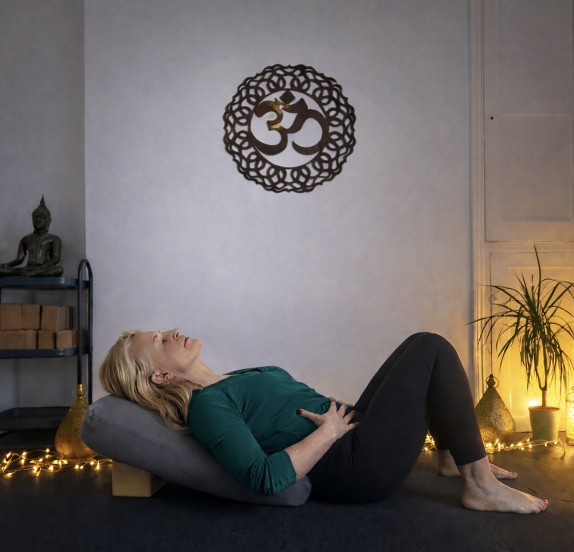 A woman lying on her back on a cushion, practicing yoga or meditation in a cozy, dimly lit room decorated with fairy lights, a Buddha statue, and spiritual symbols on the wall.