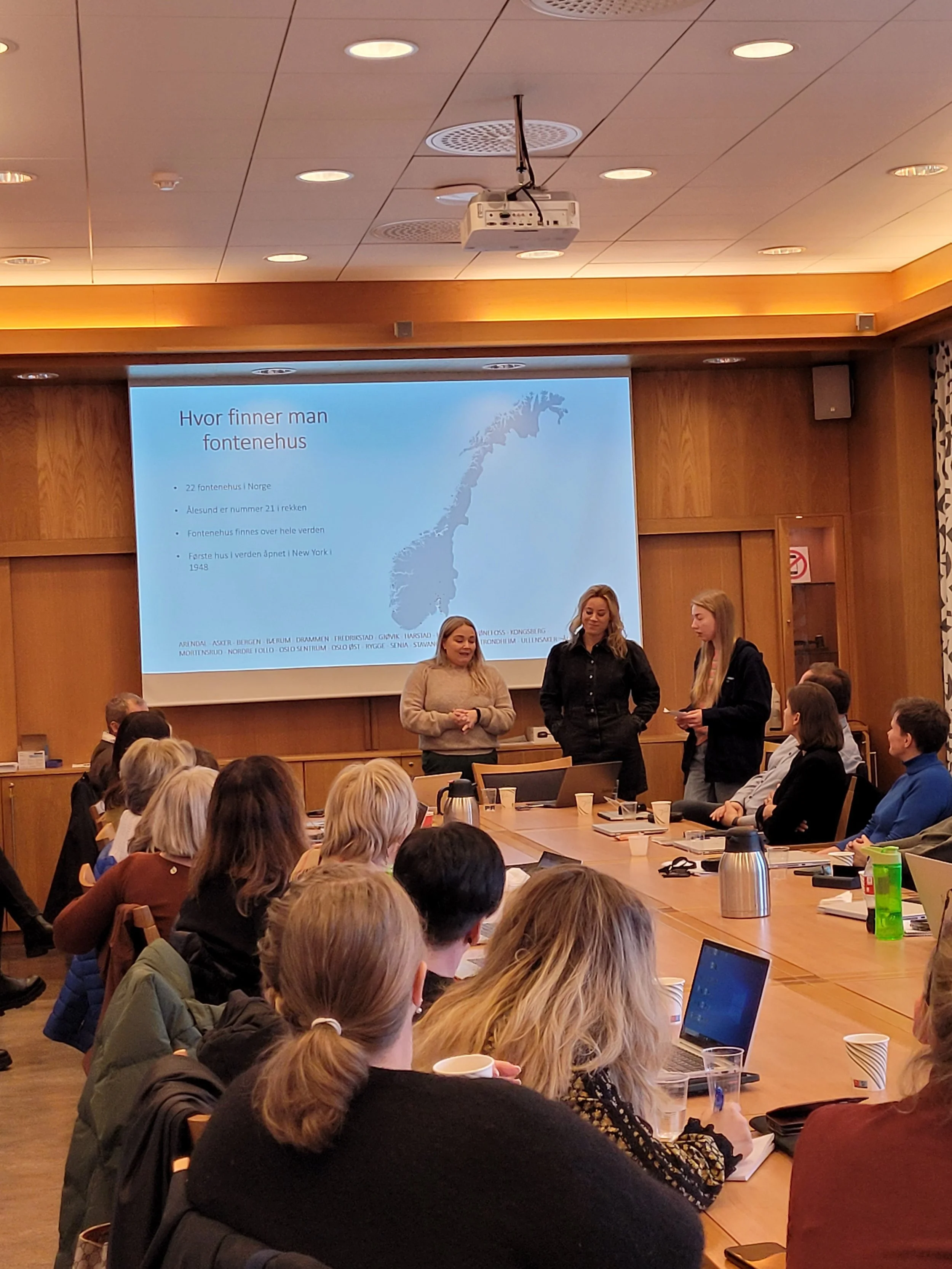 En gruppe mennesker i et møterom med en presentasjon på lerretet, hvor tre personer snakker foran forsamlingen. Presentasjonen viser en kart over Norge og teksten 'Hvor finner man fontenehus'.