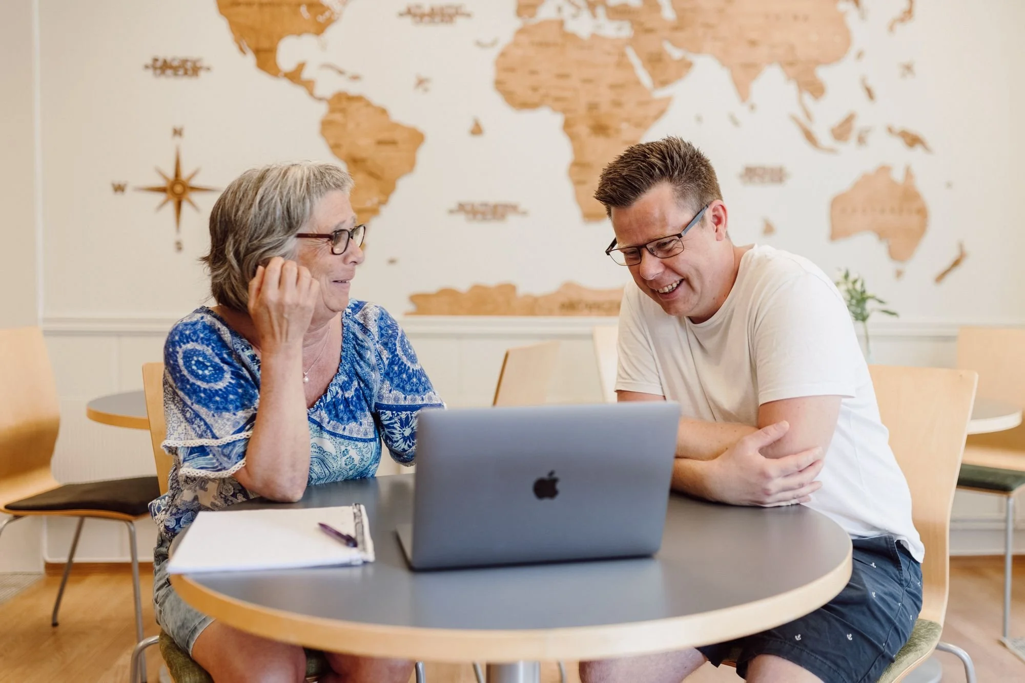 To personer som sitter ved et bord med en laptop og en notatbok, som ler og snakker i et rom med et verdenskart på veggen.