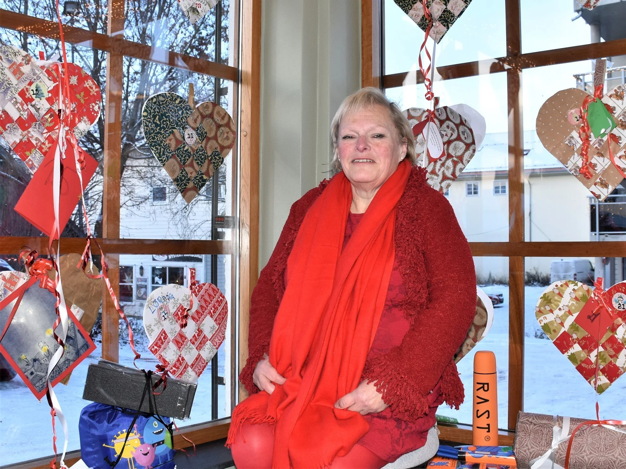 En eldre kvinne i rød genser og rød skjerf sitter foran et vindu pyntet med julehjerter og pynt, med snø og hus i bakgrunnen.