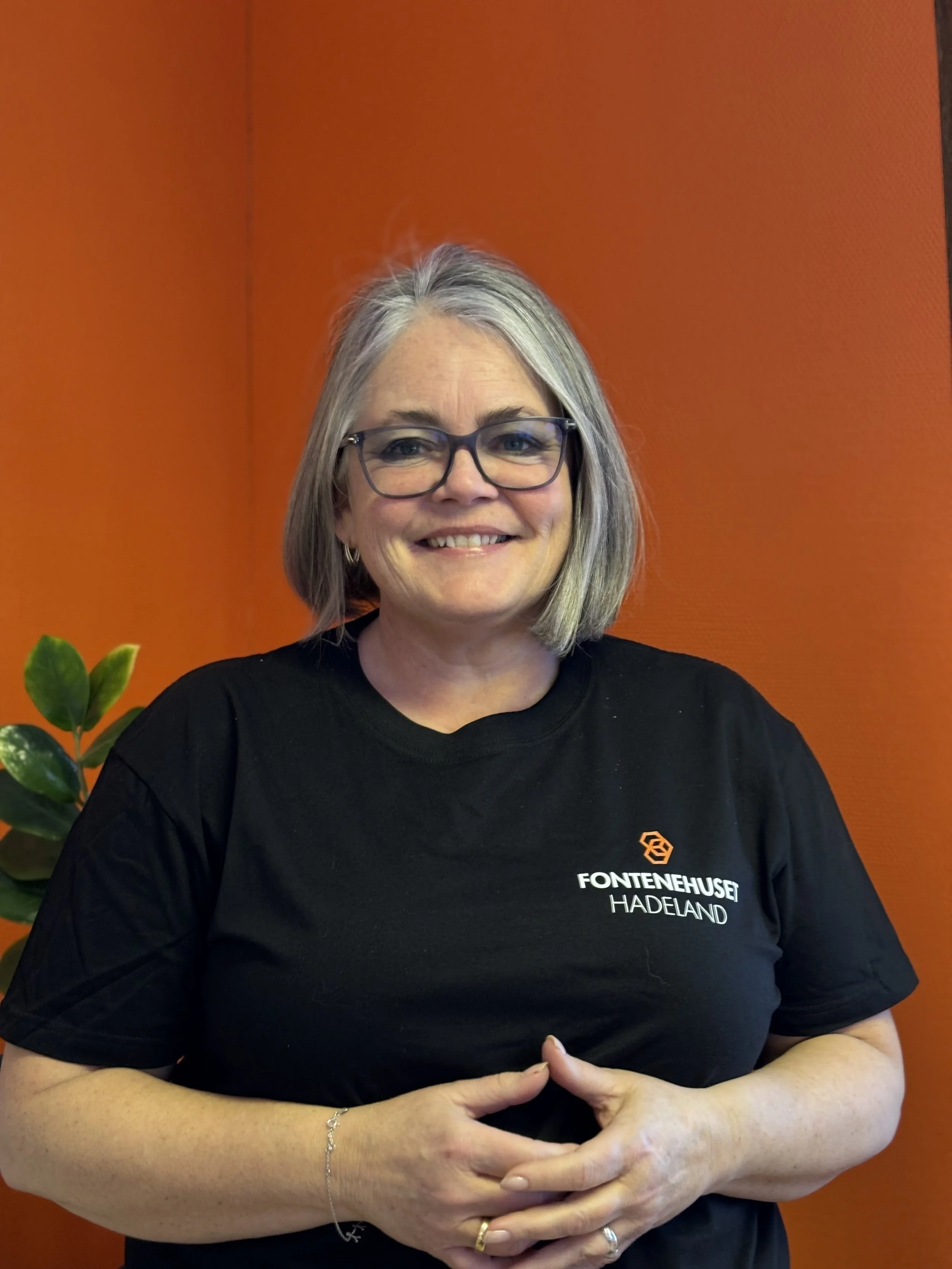 Kvinne med grått hår, briller, smilende, i svart t-skjorte med logo og tekst "FONENTEHUSET HADELAND", står foran oransje bakgrunn, med en grønn plante i bakgrunnen.