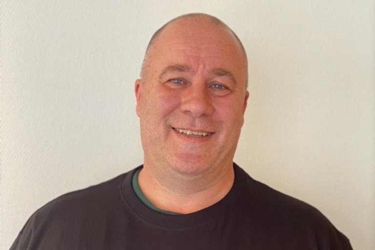 En smilende mann med kort hår og mørk T-skjorte, stående mot en lys vegg.