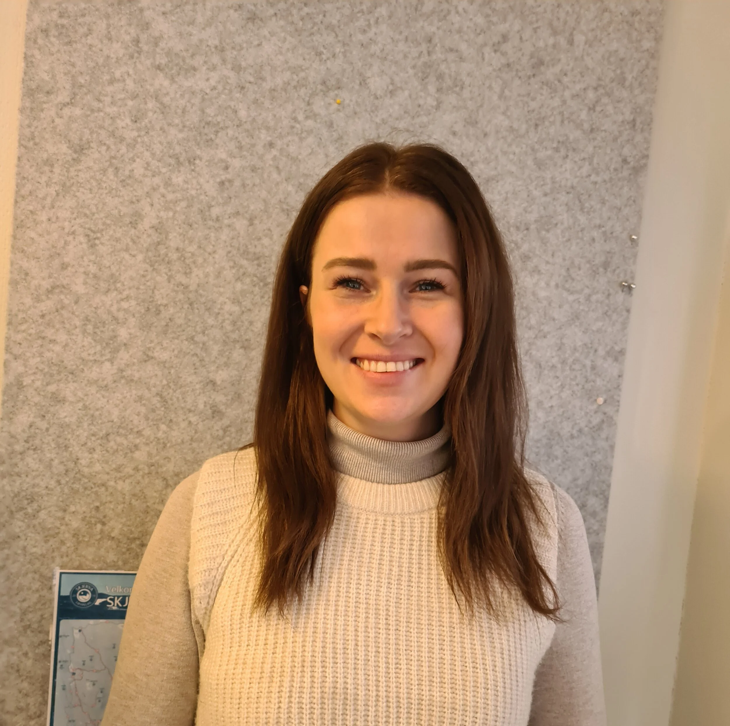 Et smilende kvinne med brunt hår, iført en beige genser med høy hals, står foran en grå vegg.