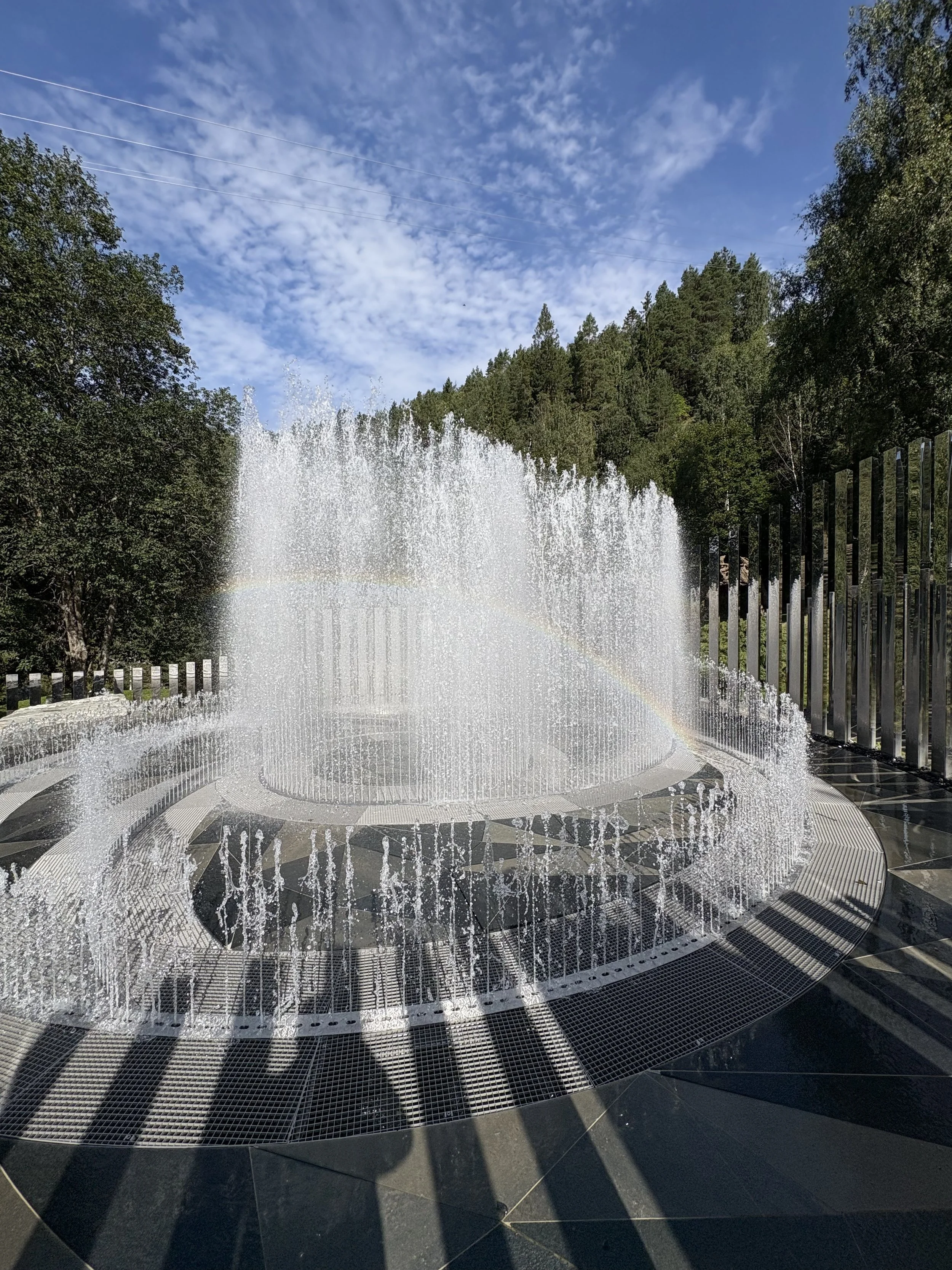 Fyrfarget vannfontene mot en bakgrunn av trær og blå himmel med skyer.