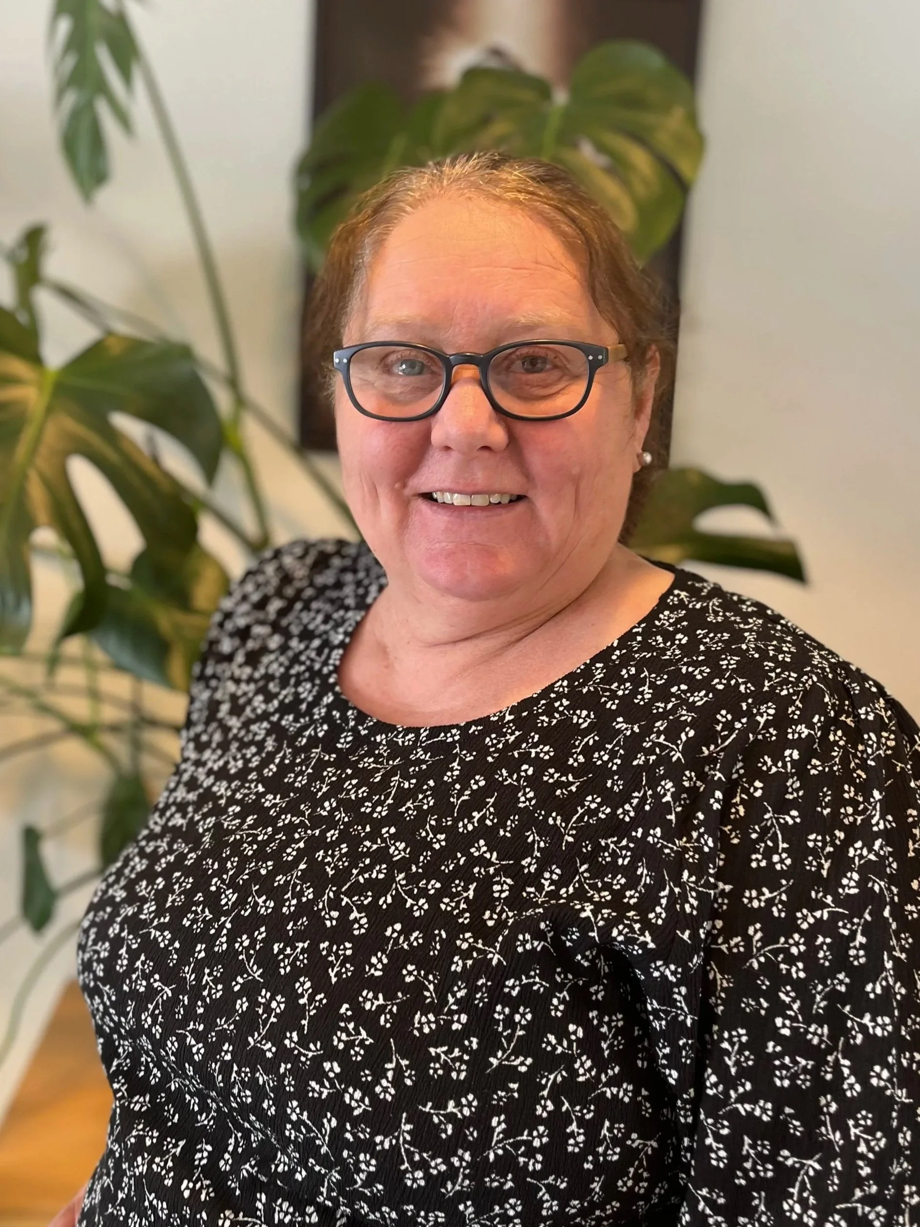 En smilende kvinne med briller og kort hår, i svart kjole med hvit blomstermønster, står foran grønne inneplanter.