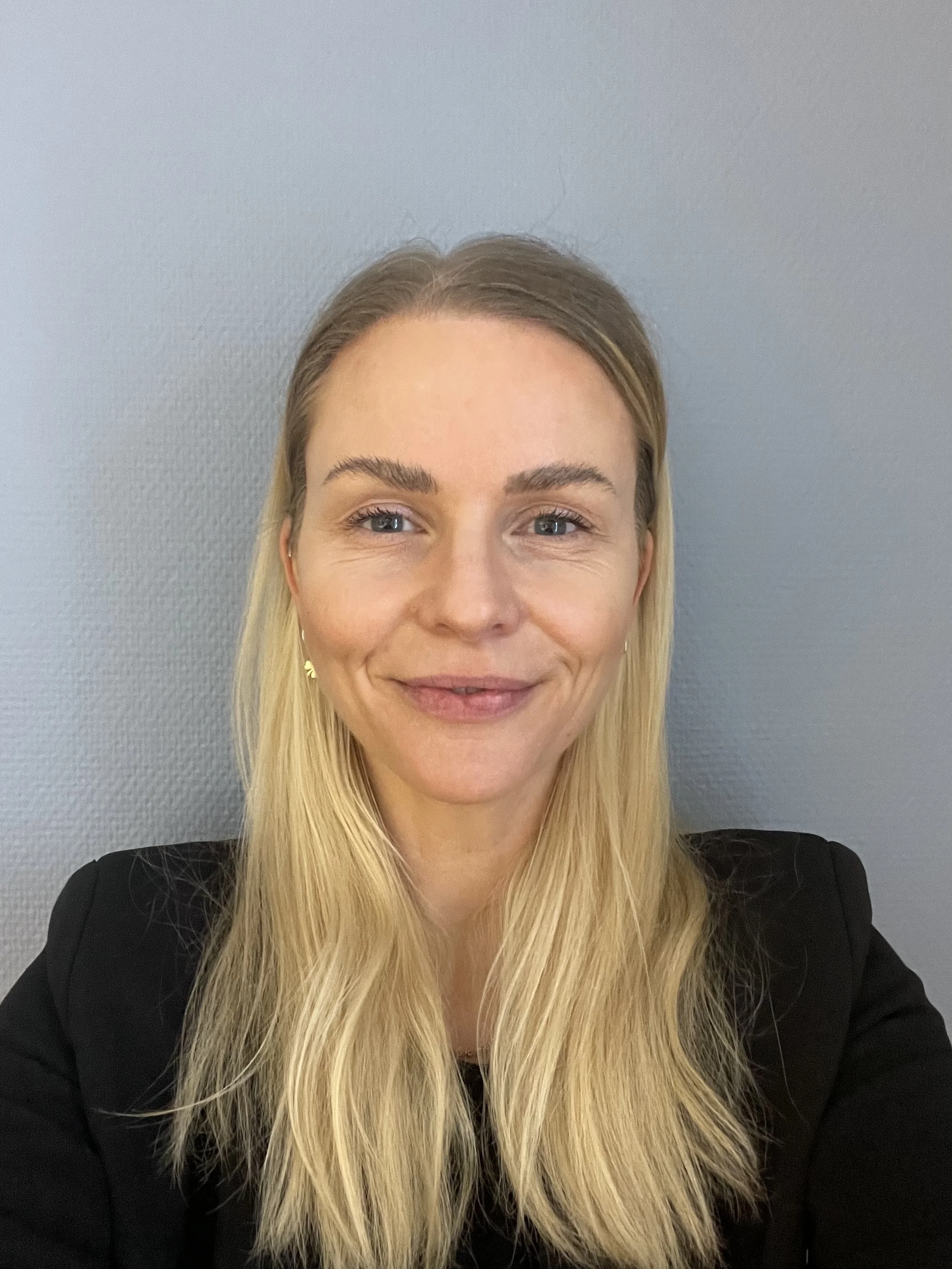 Portrett av en kvinne med blondt, rett hår, smilende, iført svart jakke, foran en grå vegg.