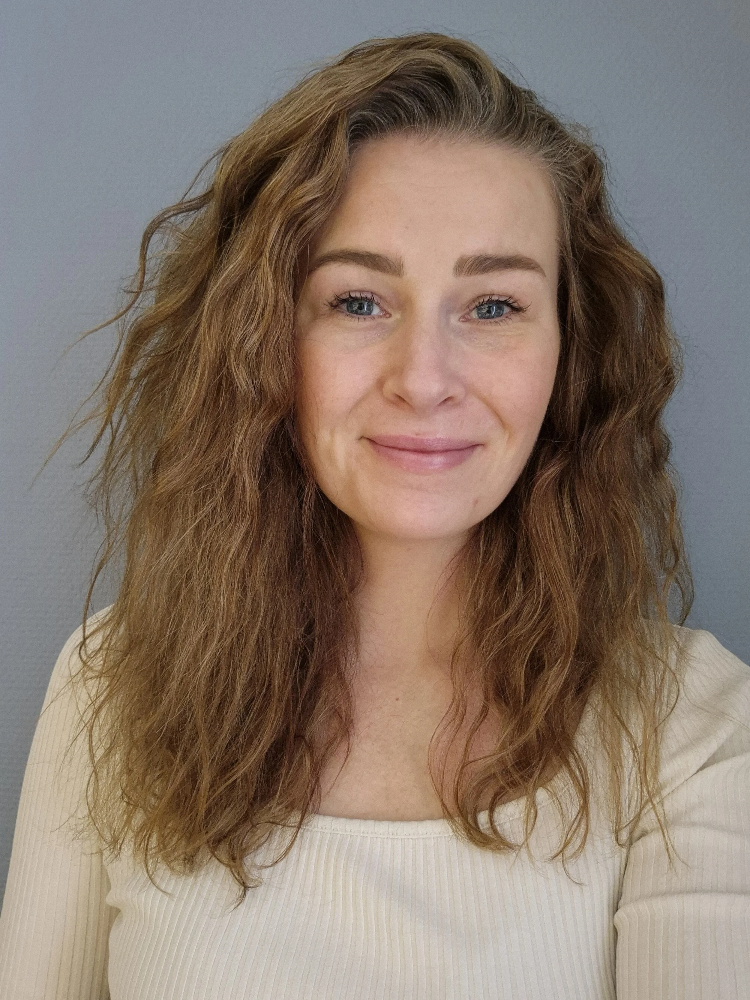 Selvportrett av en kvinne med krøllete, brunt hår som smiler lett, iført en kremfarget genser, mot en blågrå bakgrunn.