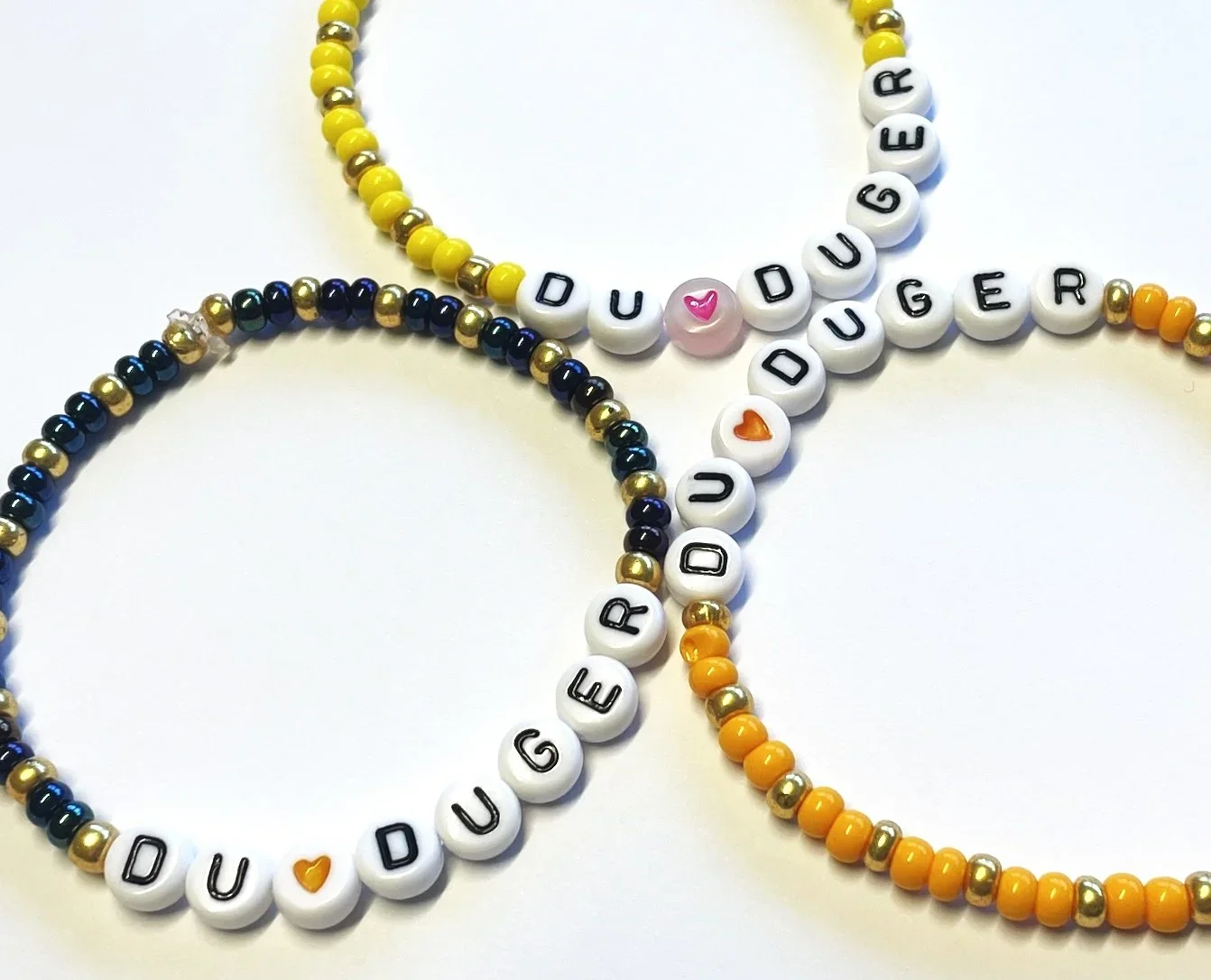 Tre perlekjede med bokstavene 'DU DUGER', dekorative perler, og små hjerteperler i forskjellige farger.