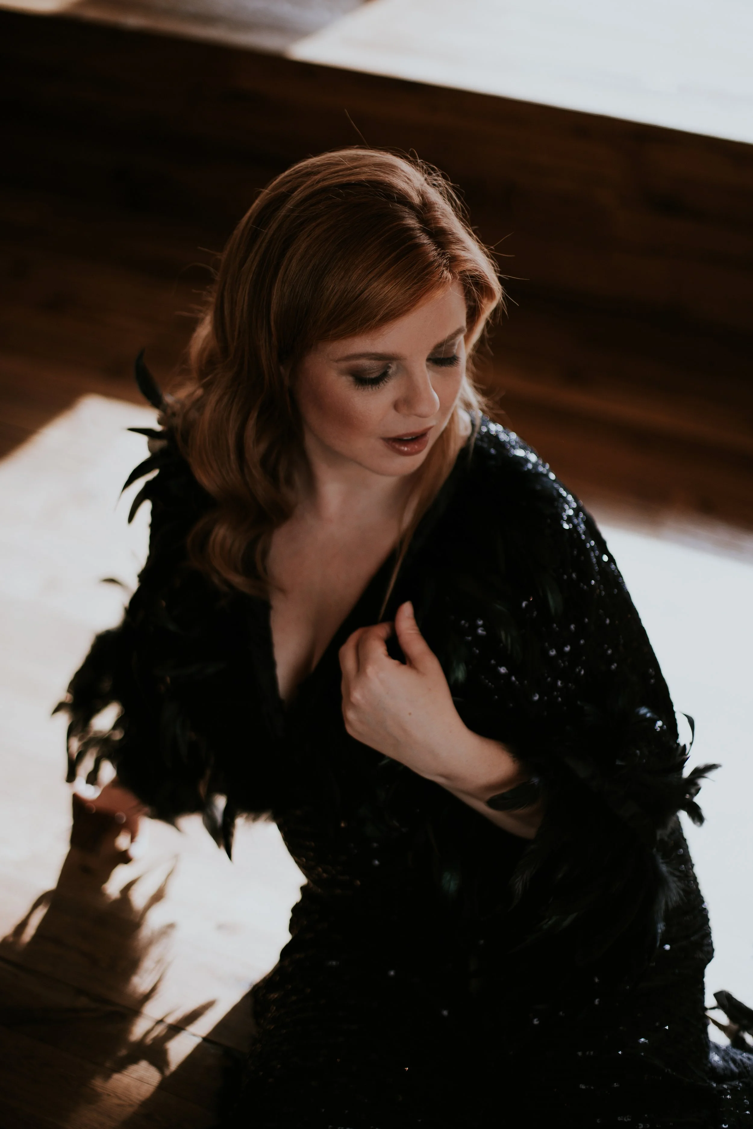 Eine Frau mit roten Haaren trägt ein schwarzes Kleid mit Federn und sitzt auf einem Holzboden, während sie nach unten blickt.