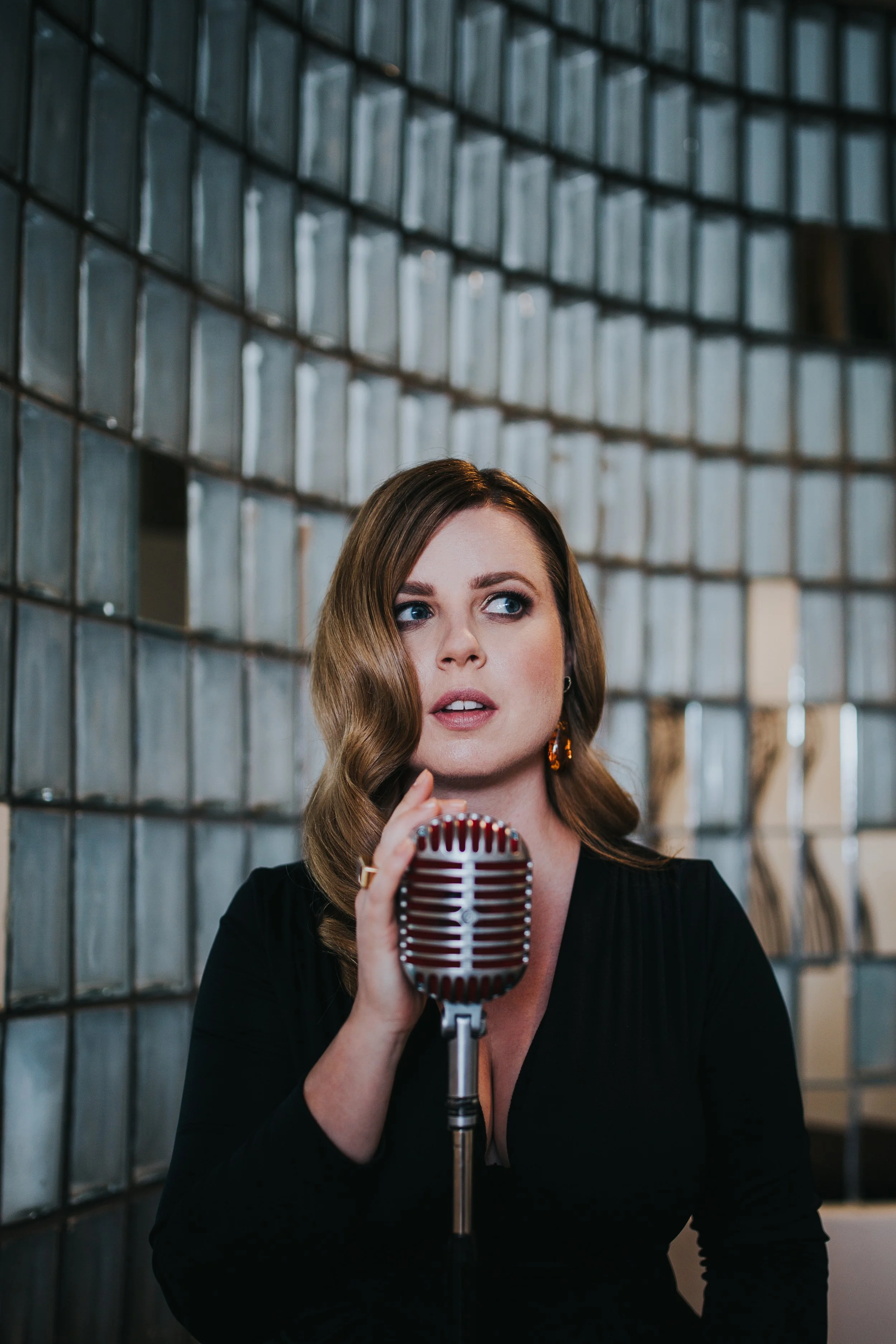 Eine Frau mit lockigem, braunem Haar, die vor einem Mikrofon steht und anscheinend singt oder spricht in einem Raum mit einer Wand aus Glassteinen im Hintergrund.