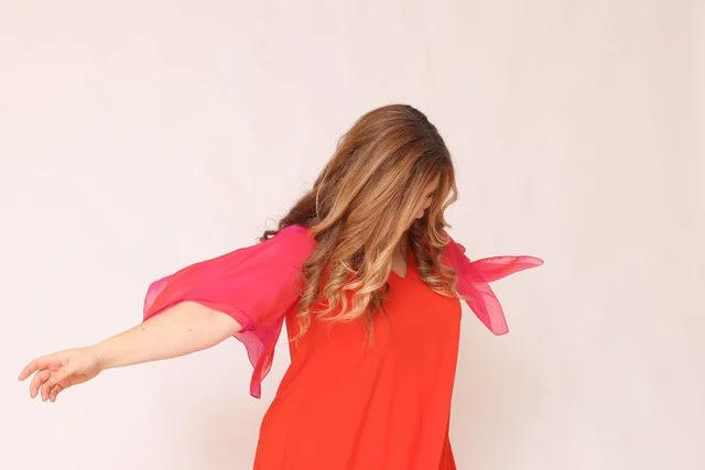 Frau in rotem Kleid mit pinken transparenten Flügelärmeln, die ihren Kopf nach unten neigt.