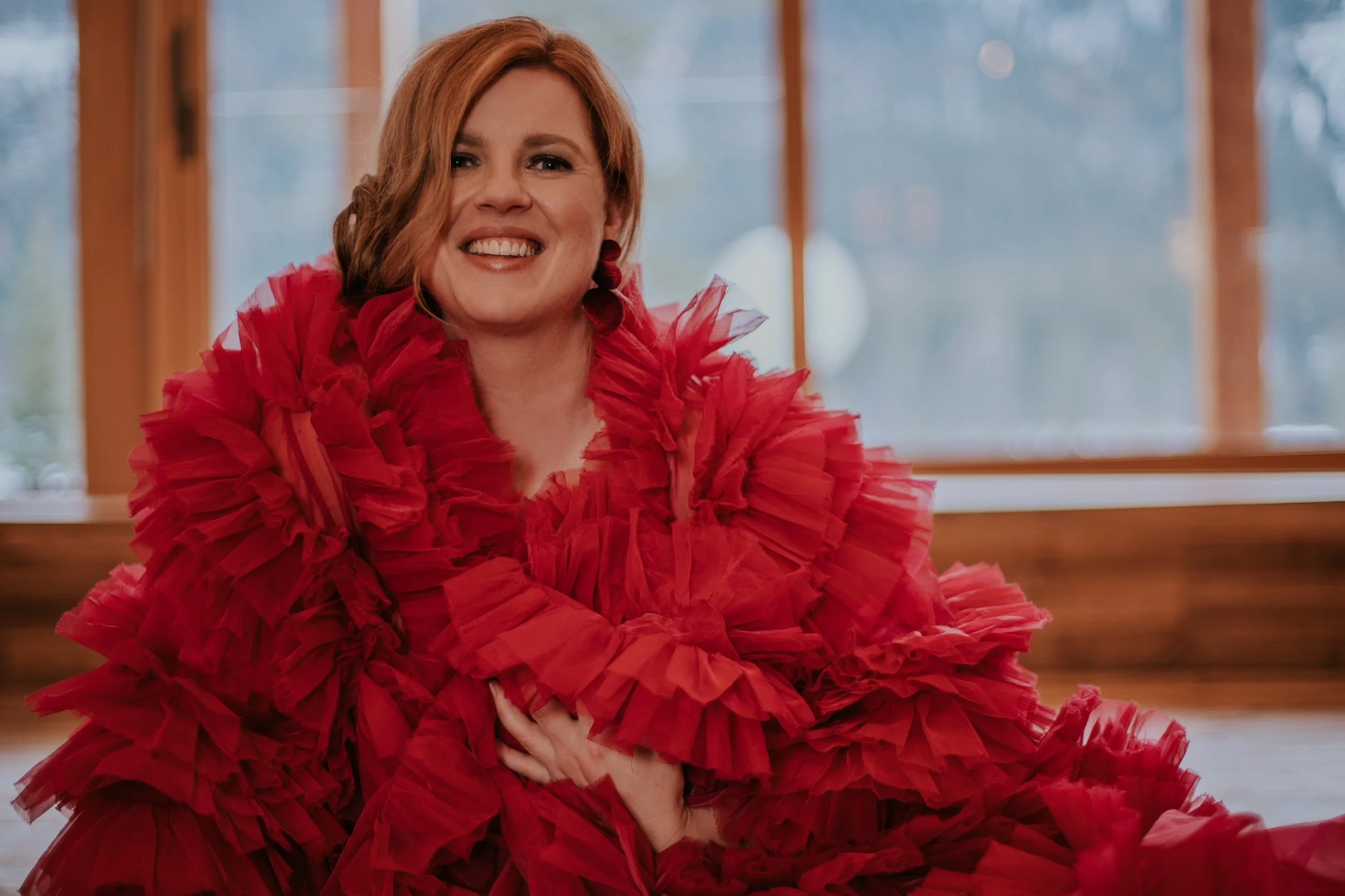 Eine Frau mit roten Haaren trägt ein rotes, rüschenreiches Kleid und lächelt fröhlich.