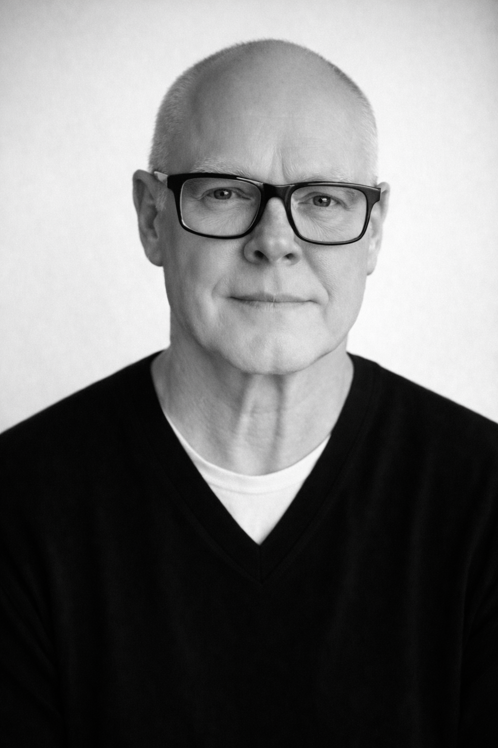 Black and white portrait of a middle-aged man with short hair, wearing glasses and a black sweater, looking at the camera with a slight smile.
