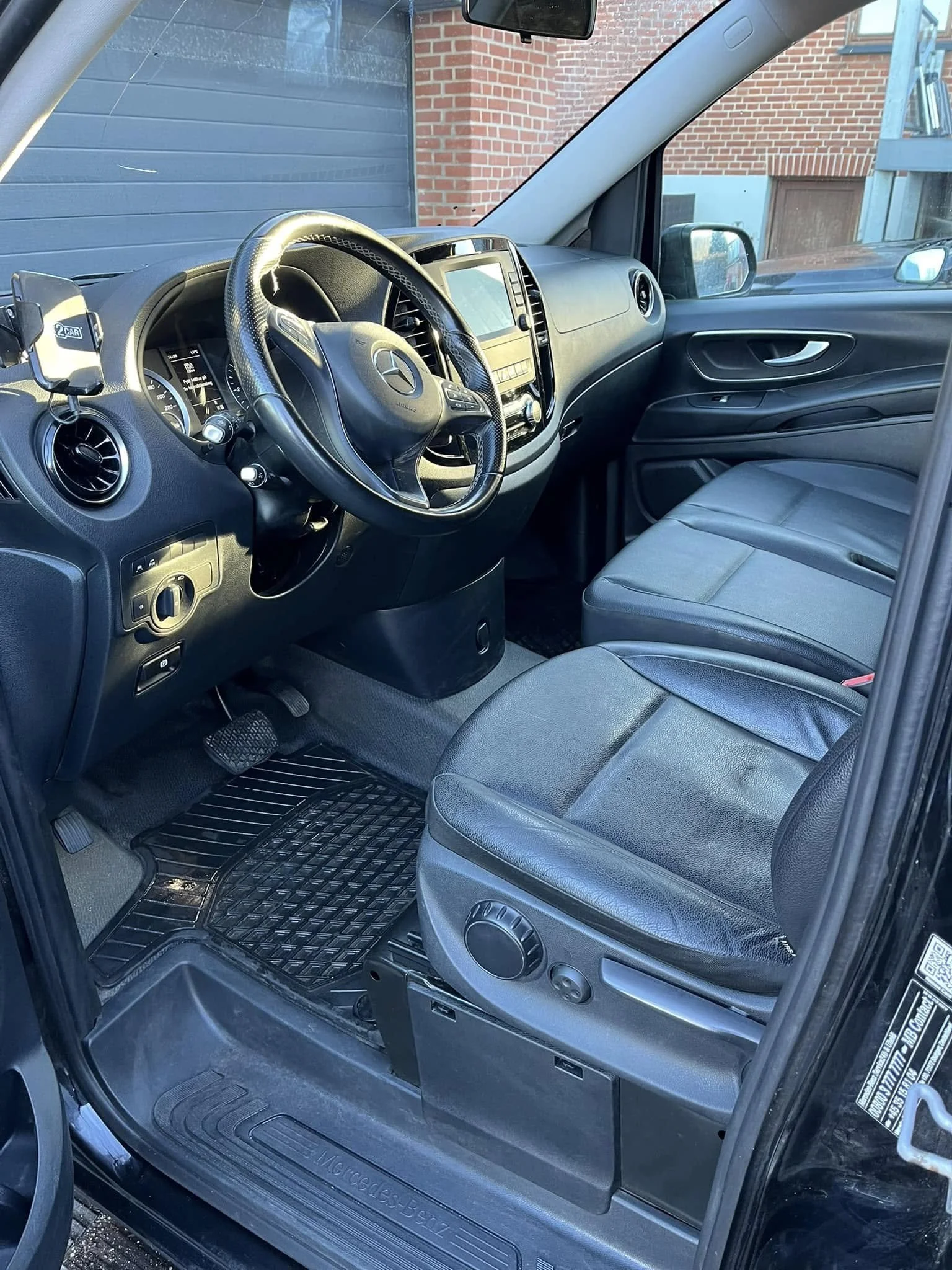 Interior of a black Mercedes-Benz van showing the driver's seat, steering wheel, dashboard with infotainment screen, and the passenger seat, with a brick building visible through the windows.