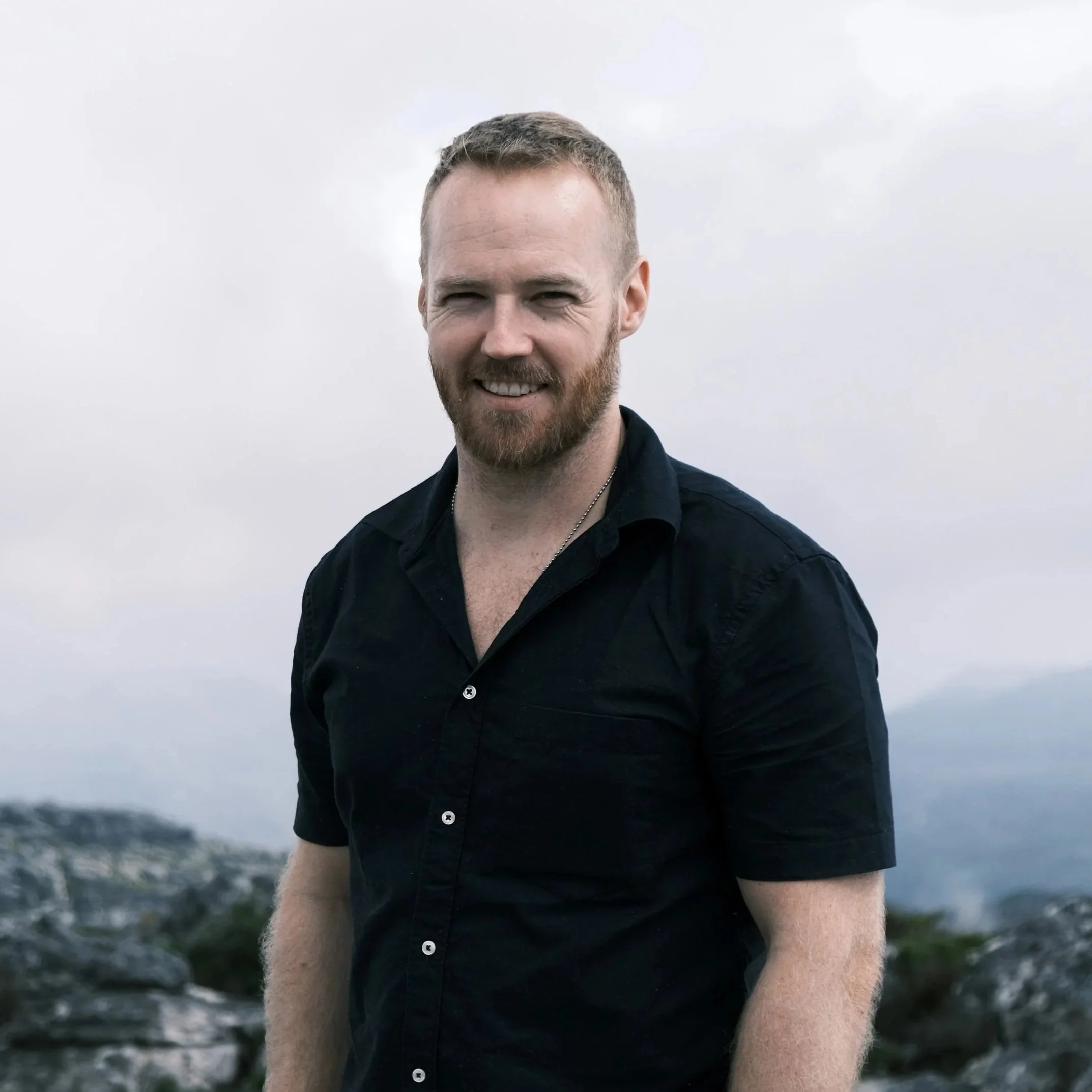 A man with short hair and a beard, wearing a dark shirt, standing outdoors on a cloudy day with mountains and rocks in the background.