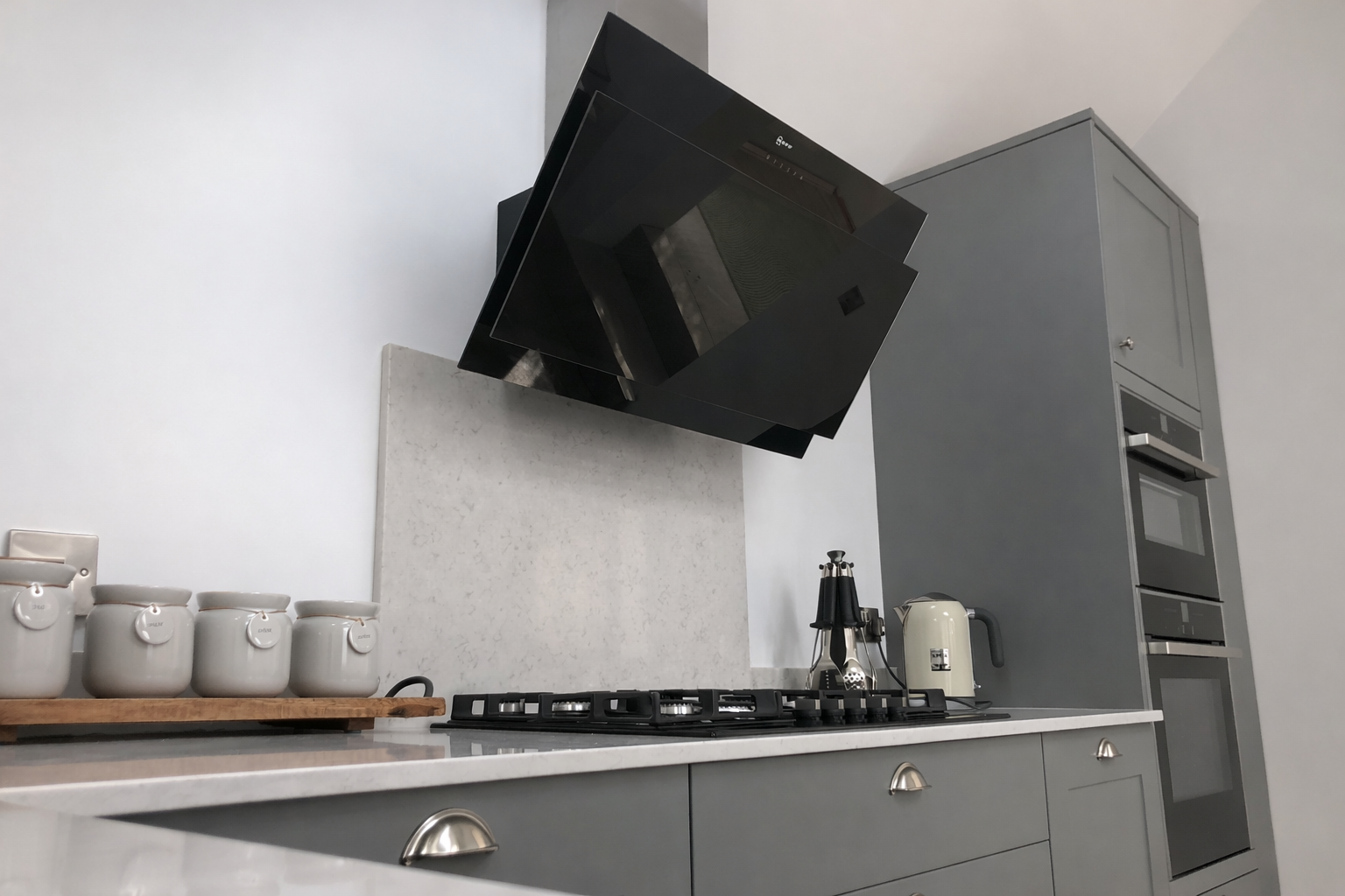 Modern kitchen with gray cabinetry, a white countertop, a black range hood above a gas stove, a wooden tray with white canisters, and small appliances including a kettle and blender.