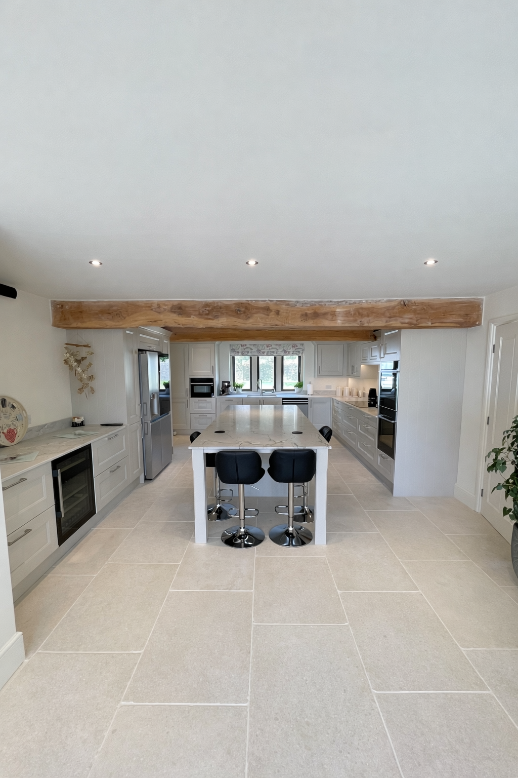 Bright, modern kitchen with white cabinets, a marble island, black bar stools, stainless steel appliances, large windows, and a wooden beam across the ceiling.