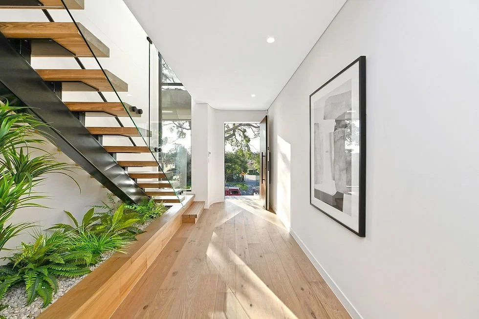Modern hallway with light-colored wooden floor, white walls, and a staircase with wooden steps and black metal support. There is a glass wall beside the staircase with plants underneath, and a large black and white framed artwork on the wall. Natural