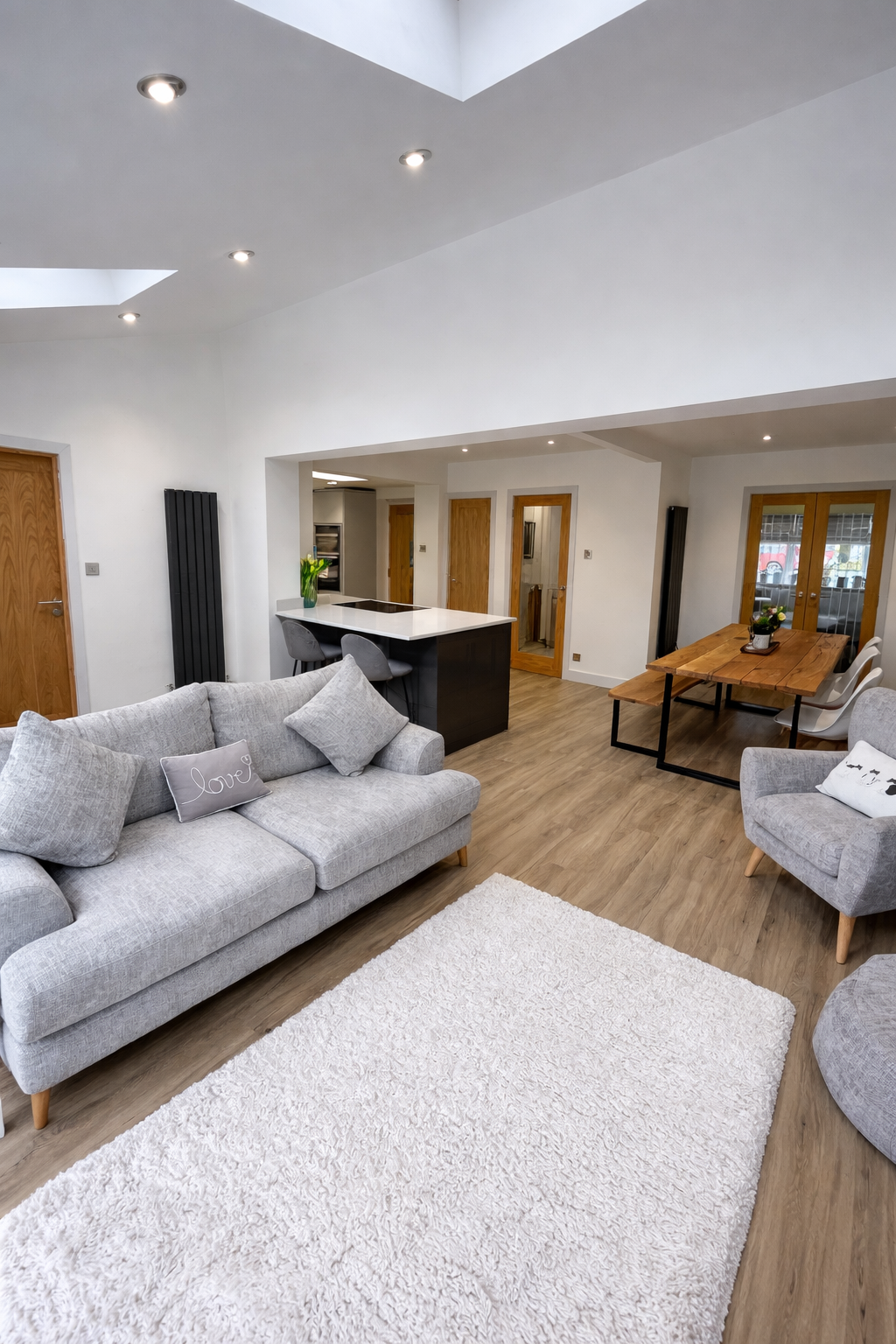A modern living room with gray sofa, armchair, white shag rug, and wooden dining table with white chairs, featuring white walls, wooden flooring, and ceiling lights.