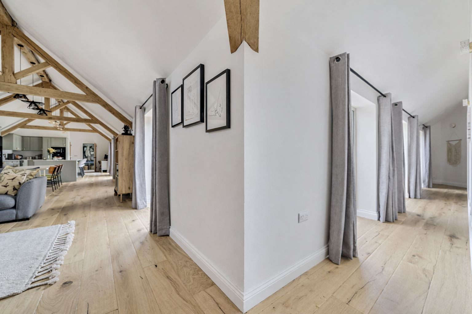 Open-plan living space with wooden floor, white walls, and exposed wooden beams on the ceiling. The area features a kitchen with gray cabinets and a dining area. Curtains are hanging on the windows along the side of the room.