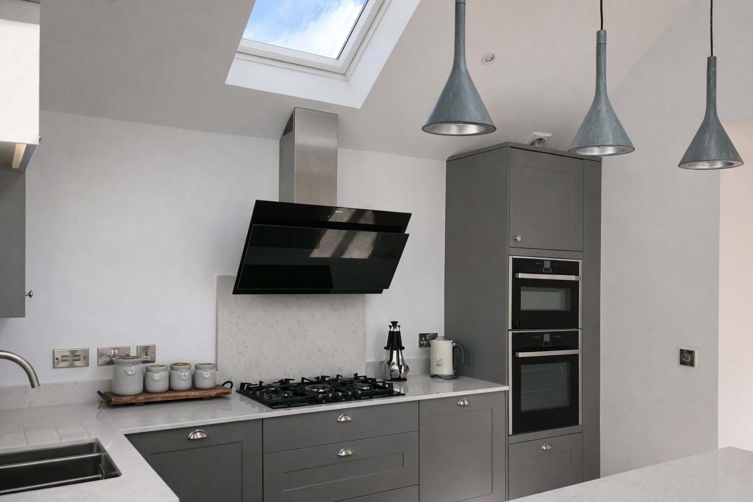 Modern kitchen with gray cabinets, black vent hood, built-in oven, skylight, and three blue pendant lights.