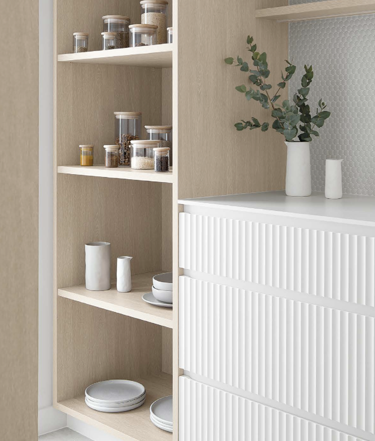 Open wooden shelves with glass jars containing spices and grains, white ceramic vases, and white dinnerware, next to a white cabinet with a textured surface, decorated with a white vase holding green eucalyptus branches and a white small vase.