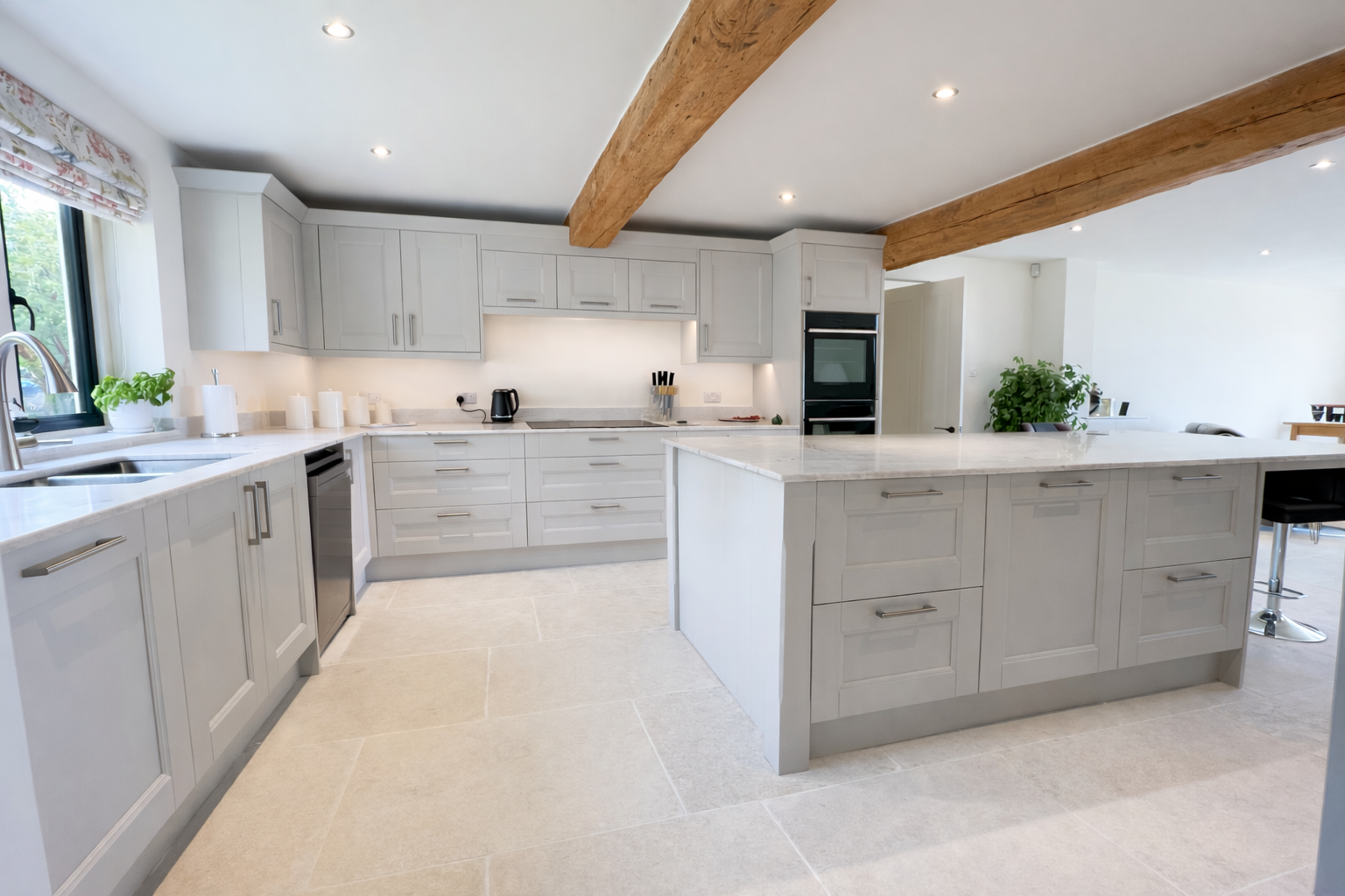 Modern kitchen with white cabinets, beige countertops, and a large kitchen island. Wooden beams are on the ceiling, and there are large beige tiles on the floor. A window with floral curtains lets in natural light.