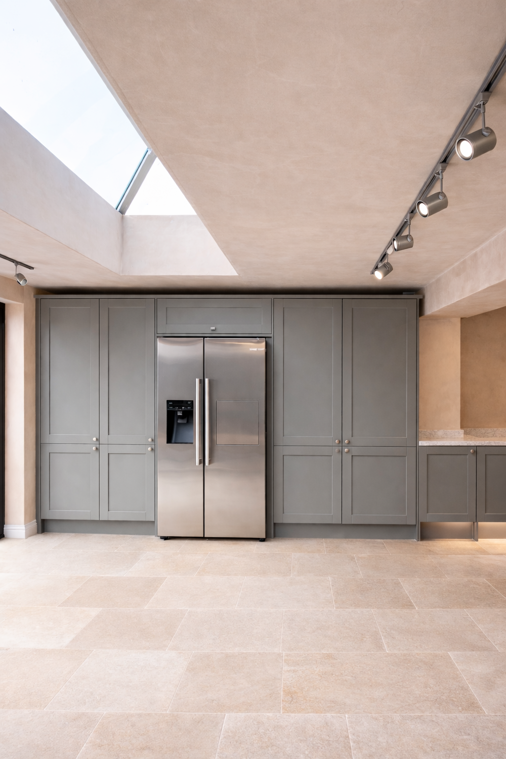 Modern kitchen with gray cabinets, stainless steel refrigerator, beige tiled floor, skylight, and track lighting.