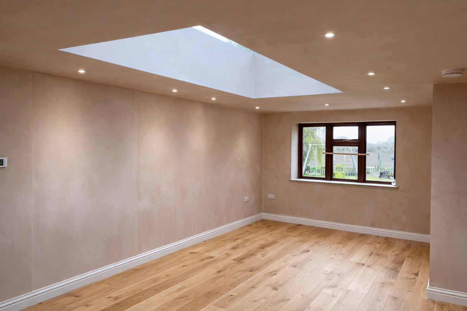 Empty room with wooden flooring, beige walls, a window showing a garden, and a skylight in the ceiling with recessed lighting.