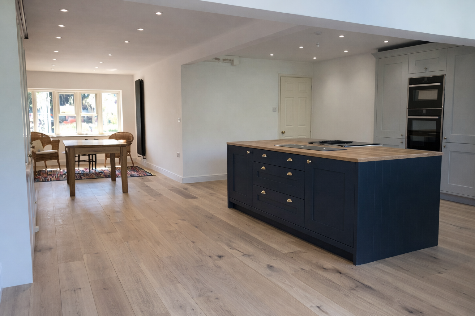 Open-concept kitchen with a navy blue kitchen island, light wood cabinets, and a light-colored wood floor. In the background, a dining area with a table, chairs, and a large window.