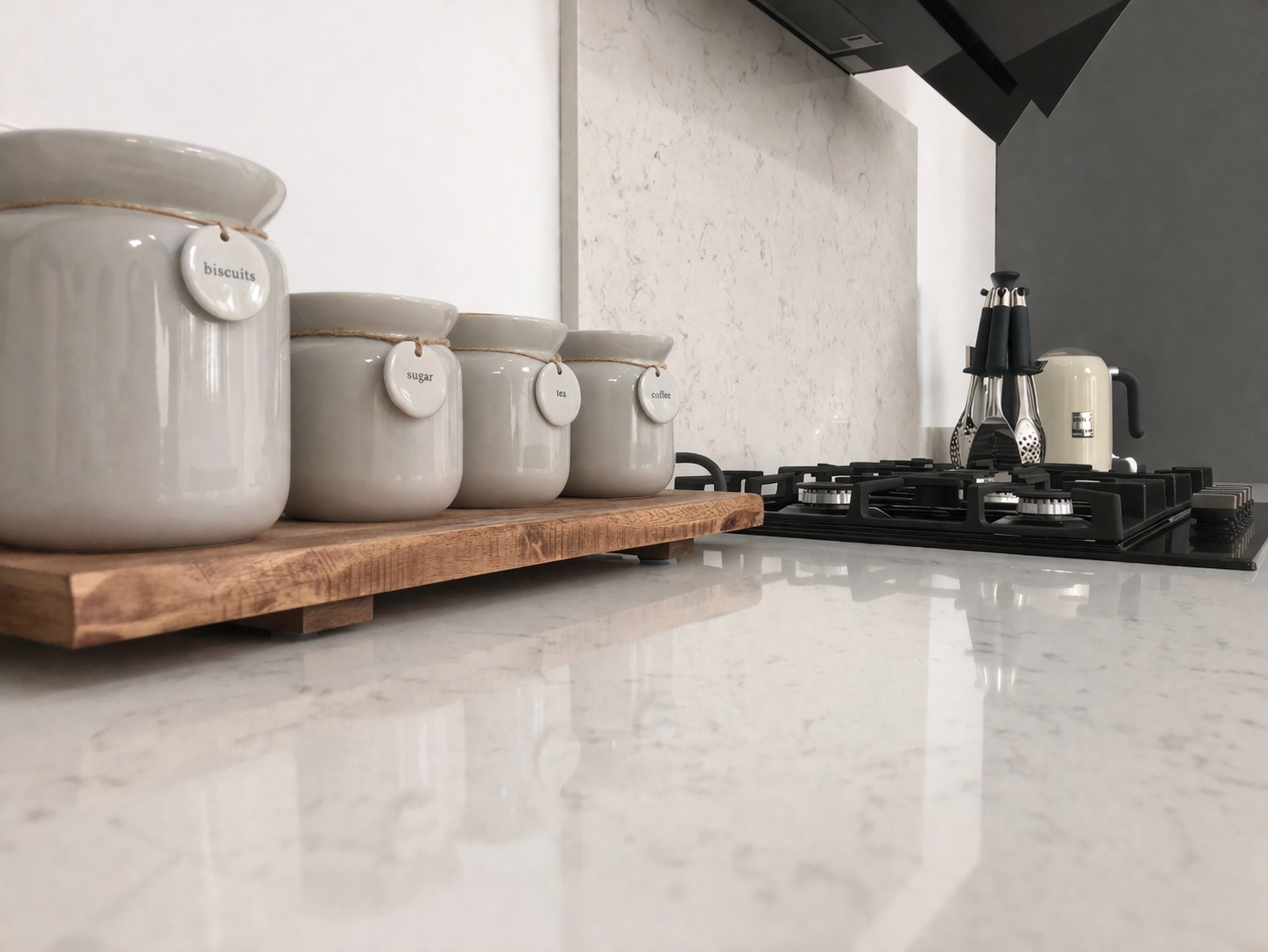 Ceramic jars labeled for biscuits, sugar, tea, and coffee on a wooden tray on white kitchen countertop, with stovetop and utensils in background.