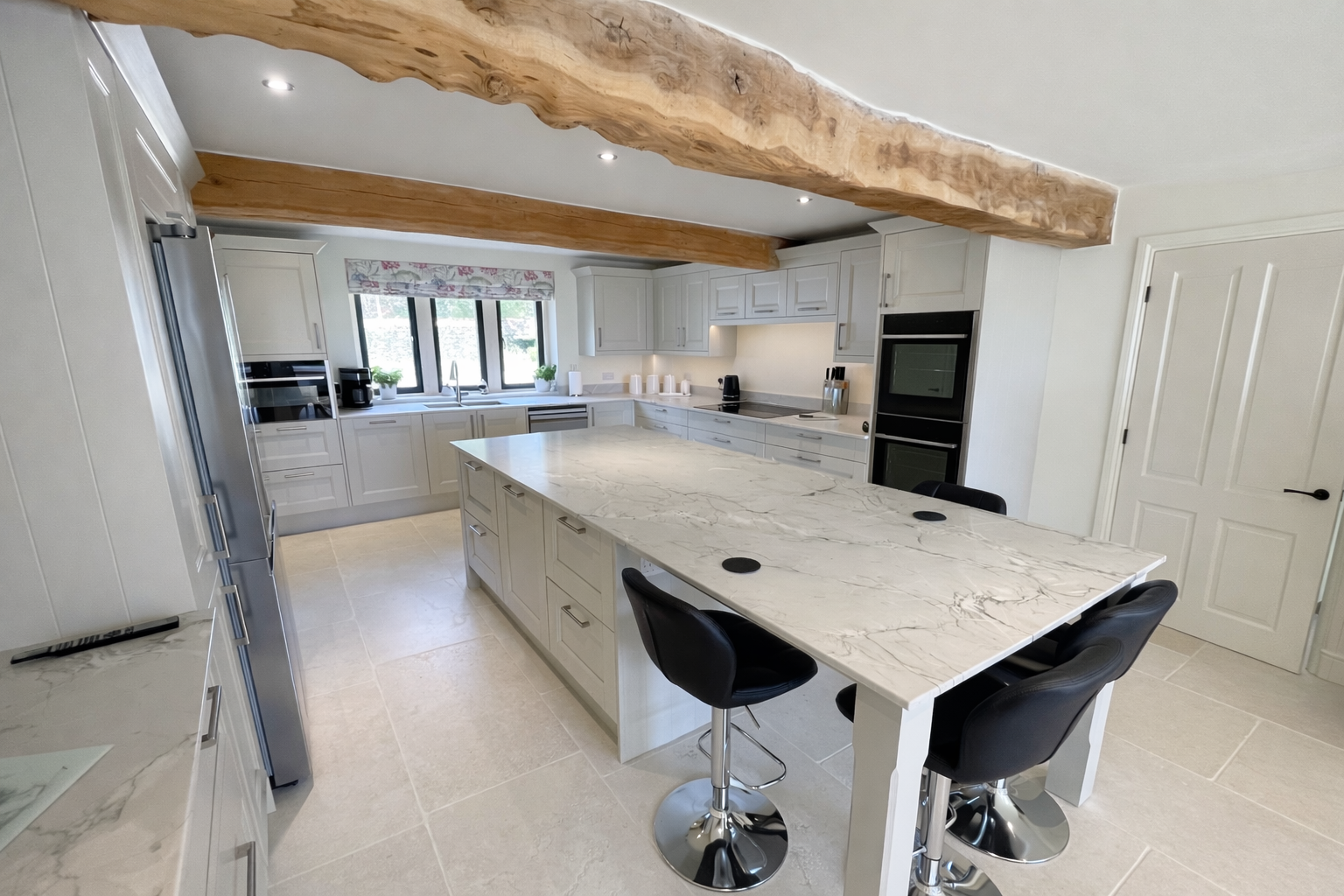 Modern kitchen with light gray cabinets, a marble island, and black barstools, featuring a large wooden beam on the ceiling and multiple windows overlooking a garden.
