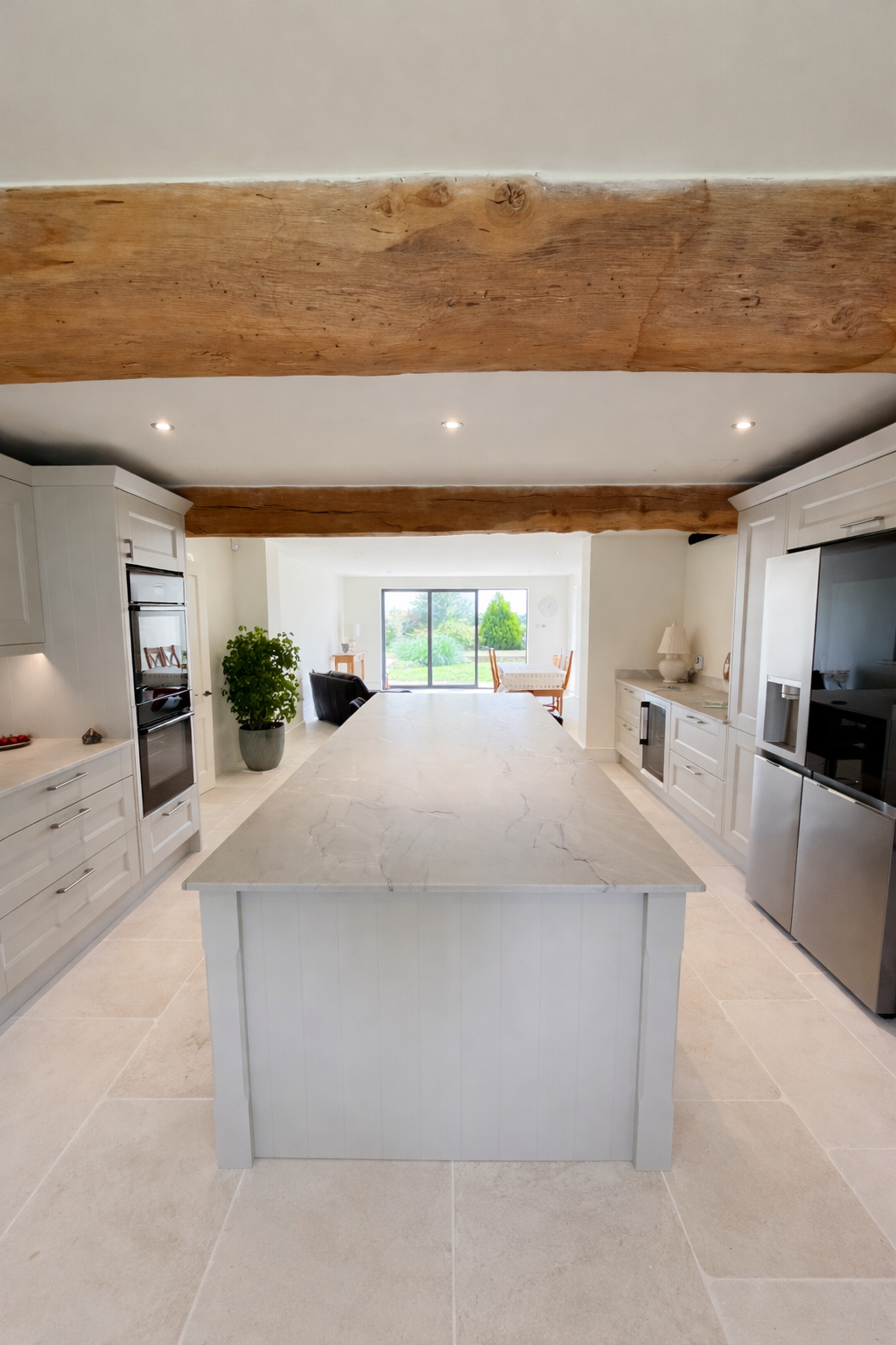 Modern kitchen with white cabinetry, marble island, stainless steel appliances, wooden beams on ceiling, and sliding glass door leading to a garden.