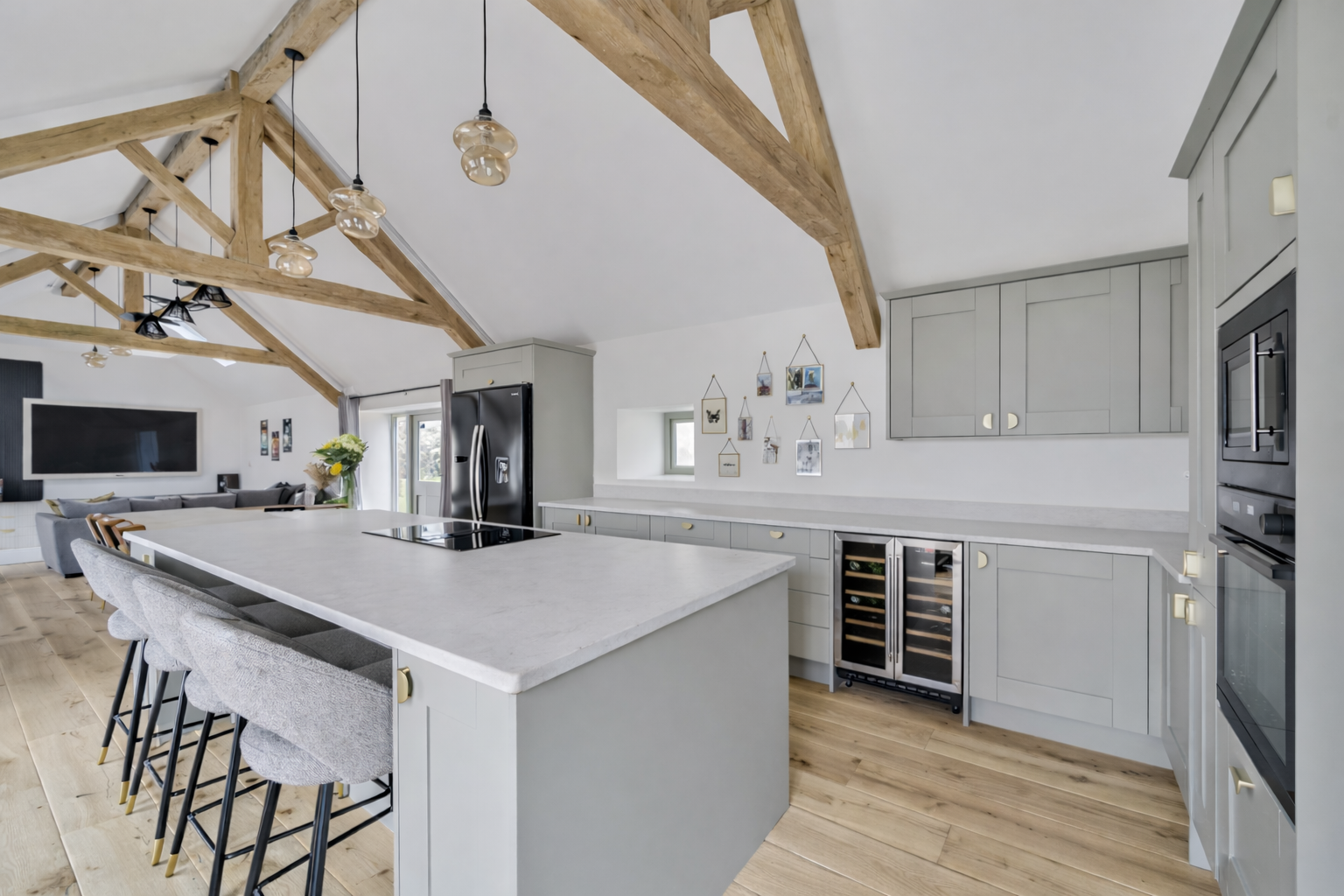 Modern kitchen with light gray cabinets, an island with bar stools, a black refrigerator, built-in oven, wine cooler, and wood beams on the ceiling. In the background, there is a living area with a large TV and a gray sofa.